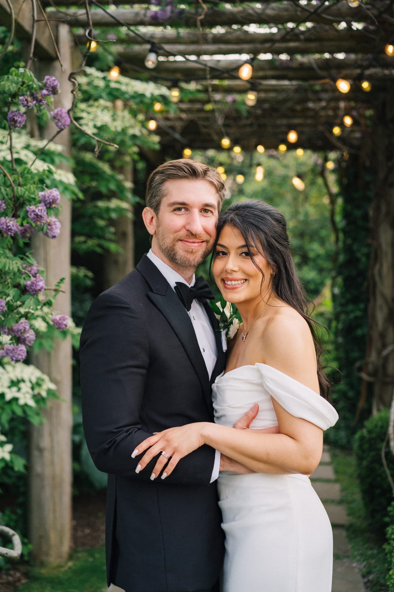 A couple stands together in a garden, the bride in a white dress and the groom in a black suit, surrounded by purple flowers and string lights.