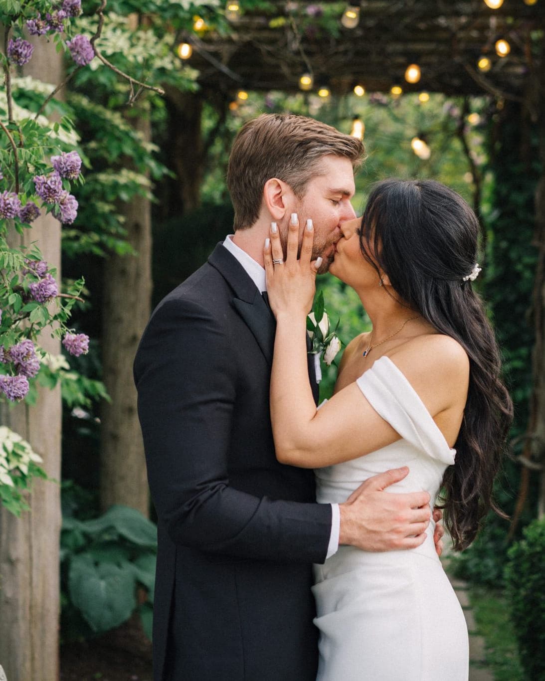 A couple in wedding attire shares a kiss under a trellis adorned with flowers and string lights.