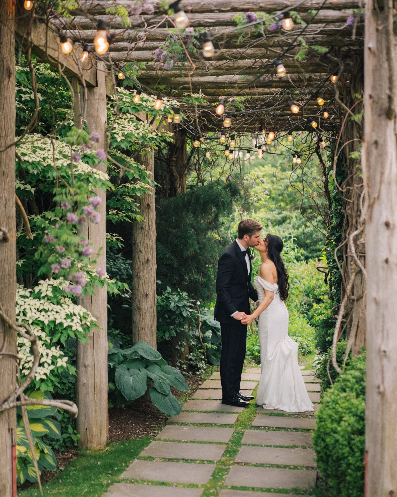 A bride in a white gown and a groom in a black suit kiss under a wooden pergola adorned with string lights and greenery.