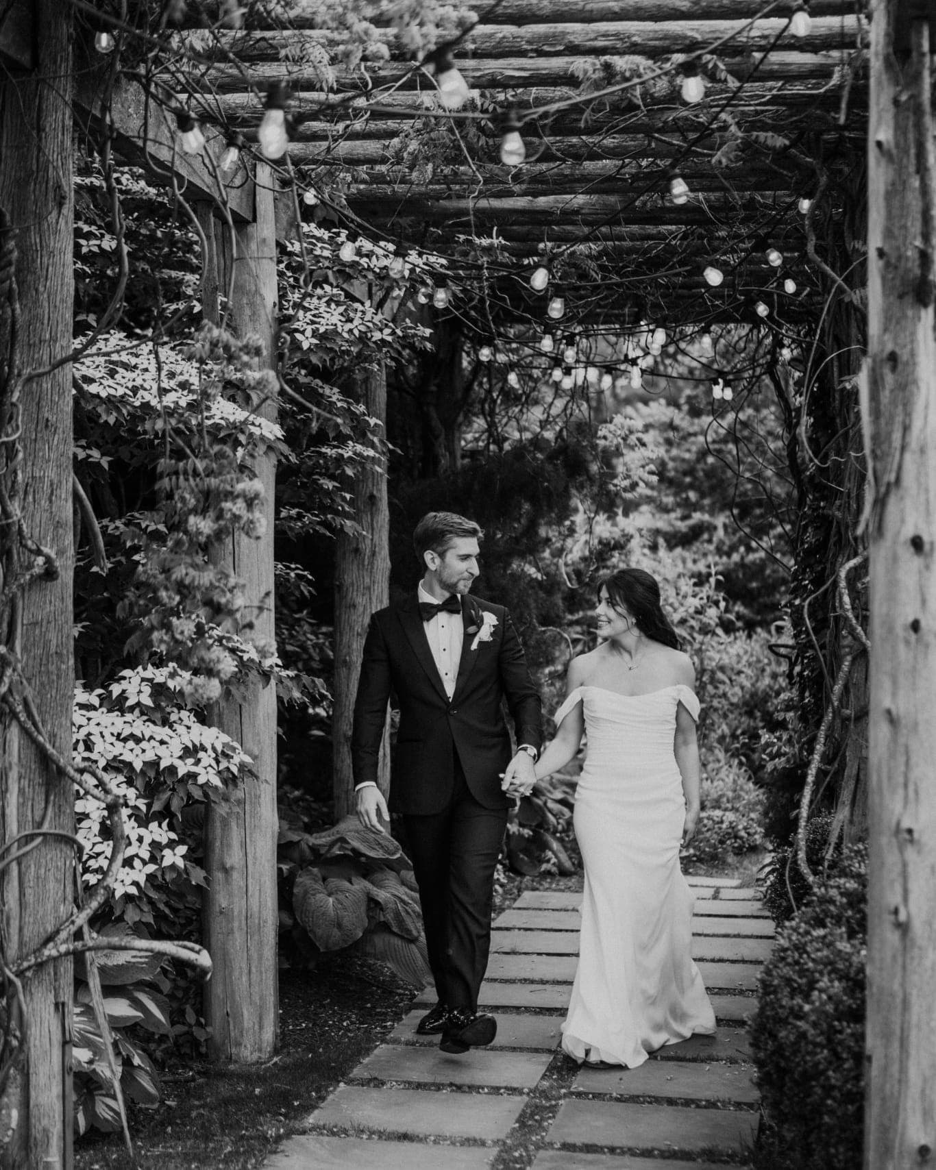 A bride and groom walk hand in hand under a wooden pergola adorned with string lights.