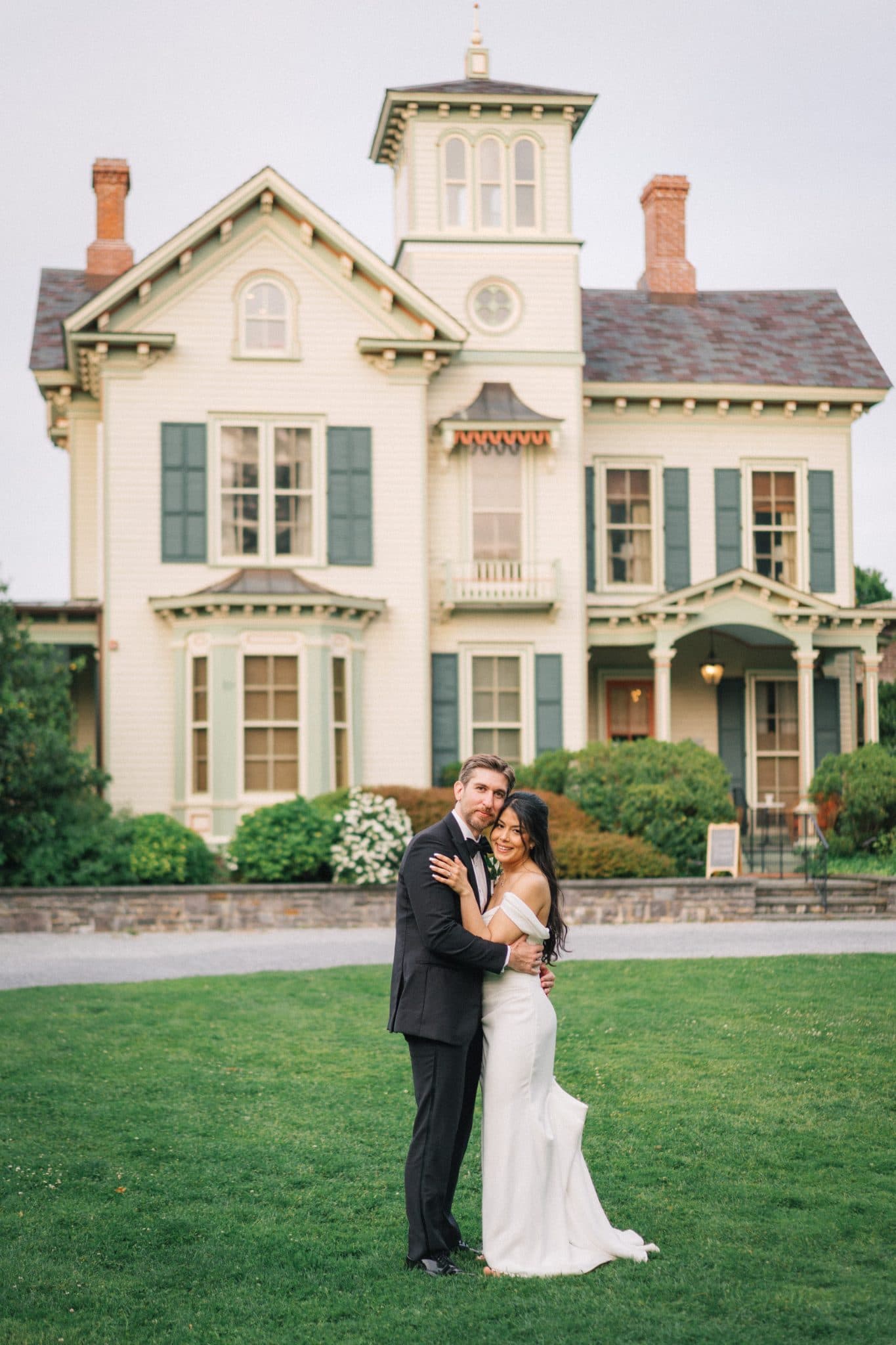 A couple stands in front of a large, ornate house with green shutters and a grassy lawn.