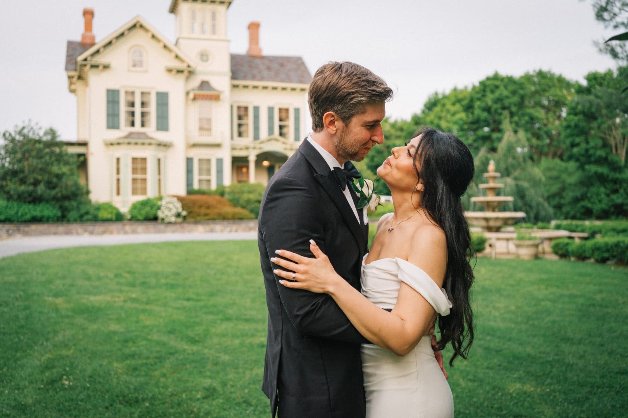 A bride and groom embrace in front of a large, white house with green shutters and a fountain in the background.