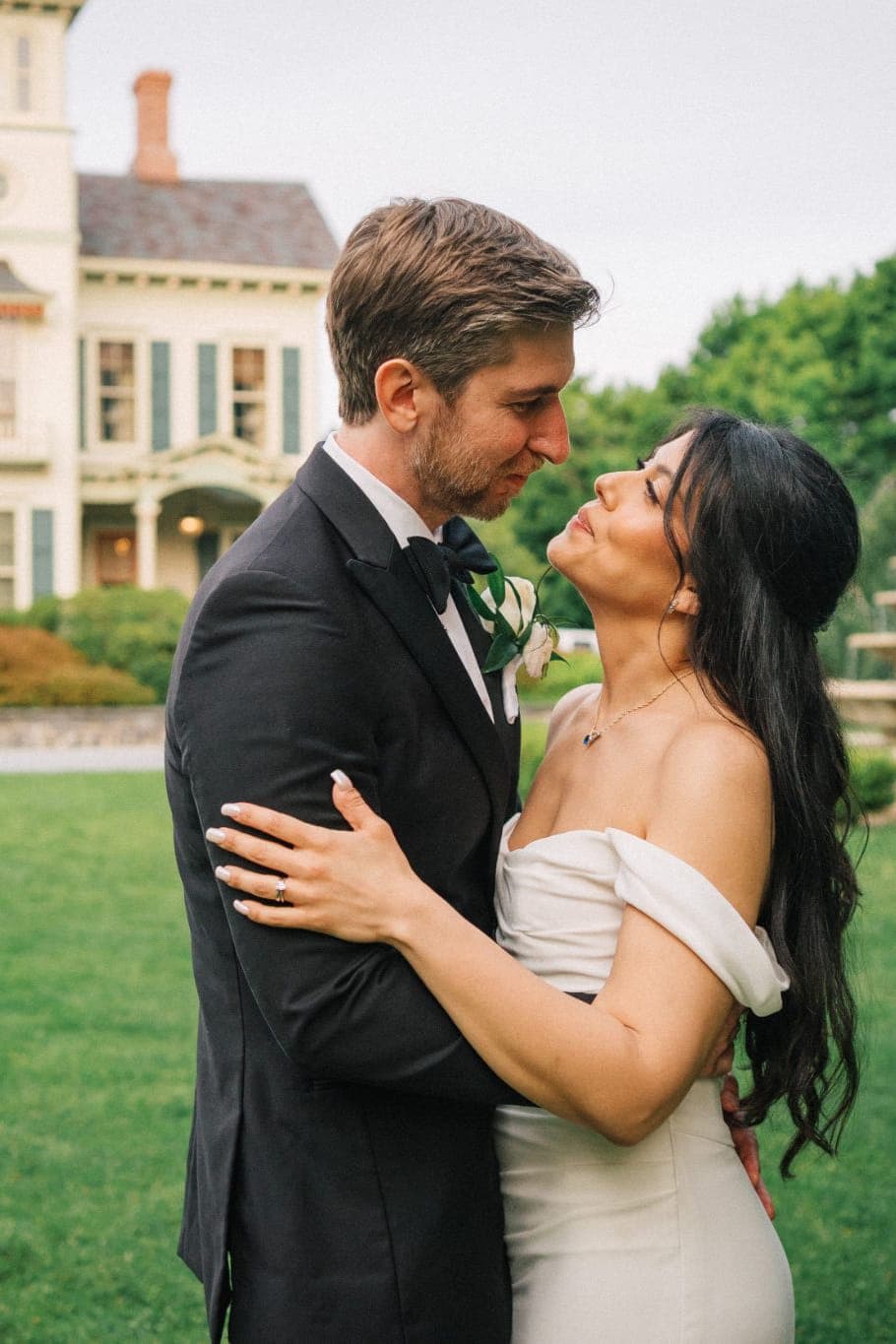 A bride and groom embrace in front of a large, white house with green shutters and a fountain in the background.