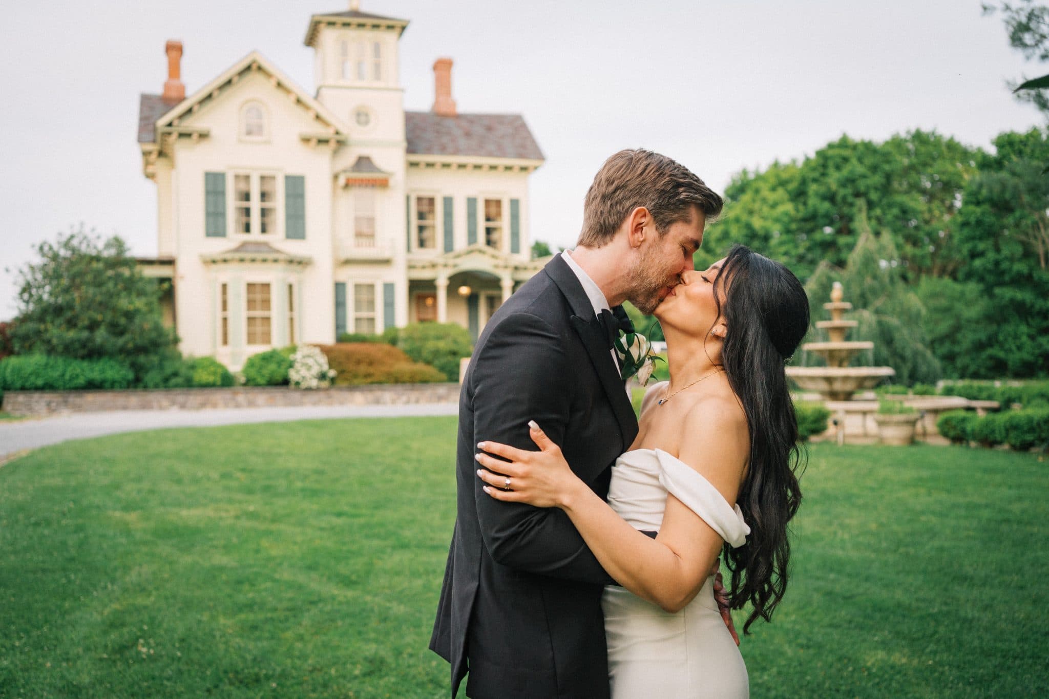 A bride and groom share a kiss in front of a large, elegant house with a fountain and lush greenery.