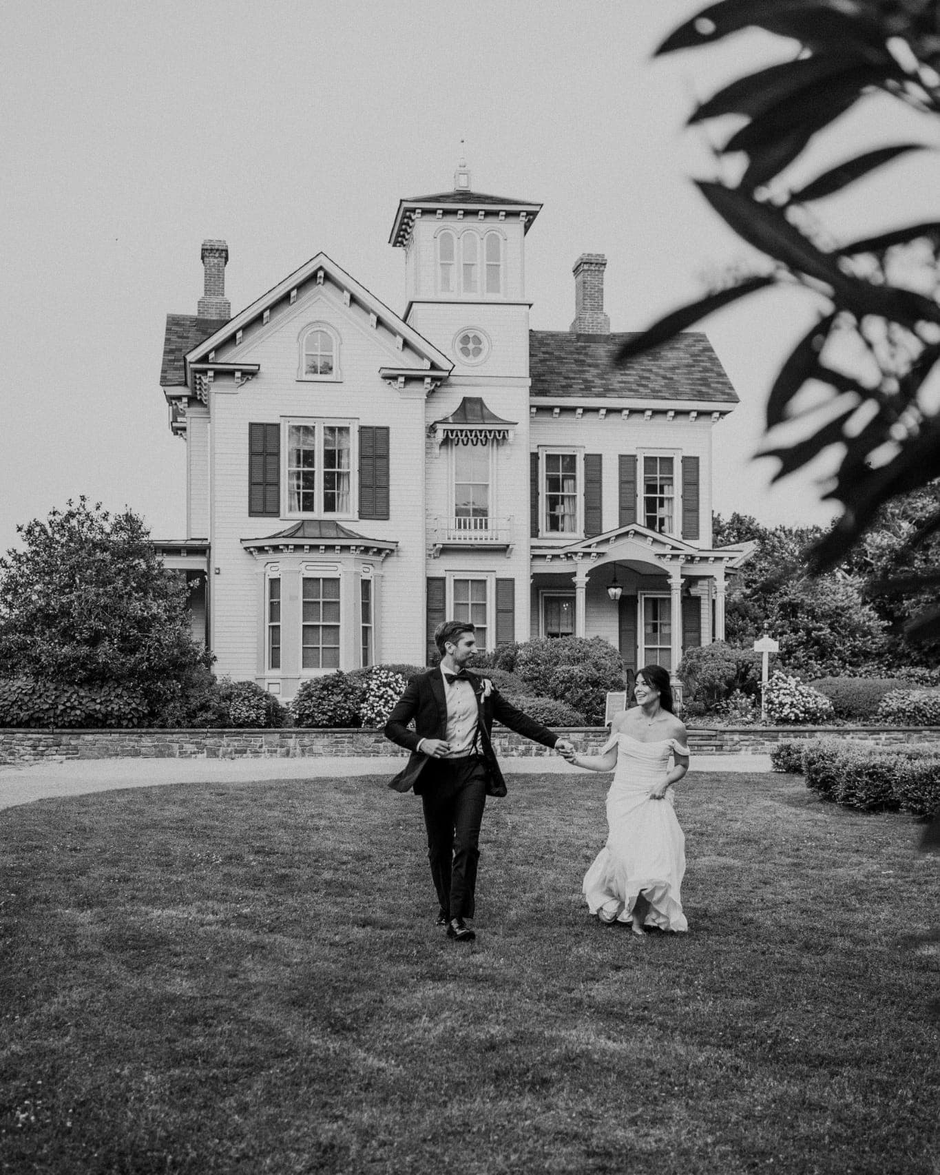 A couple in formal attire walks hand in hand across a grassy lawn in front of a large, multi-story house.