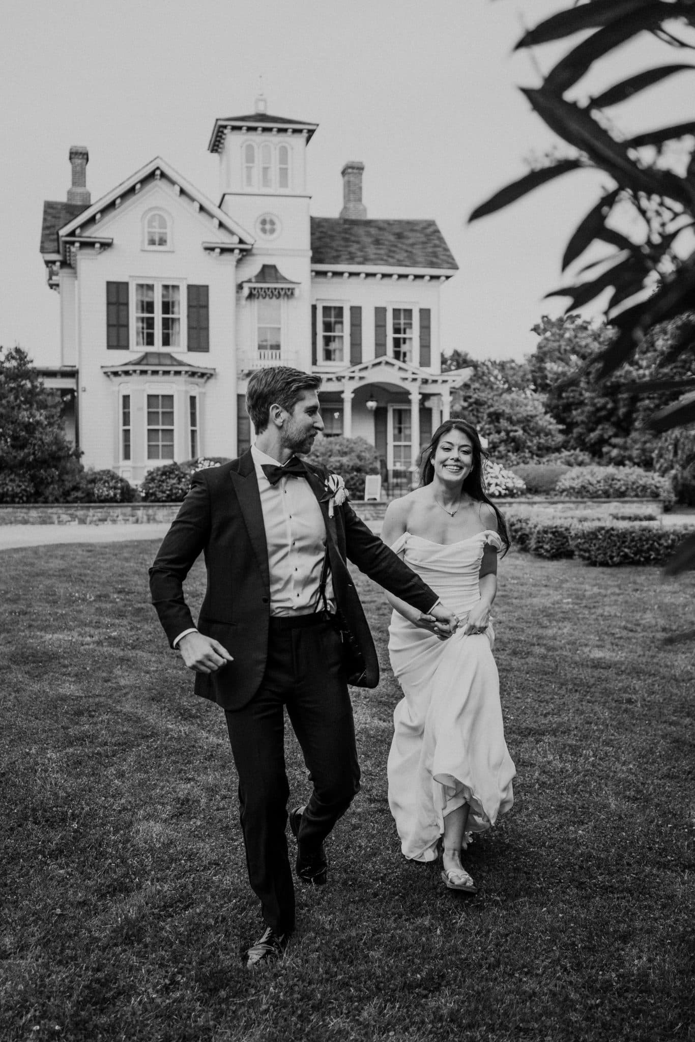 A bride and groom walk hand in hand in front of a large, historic house.