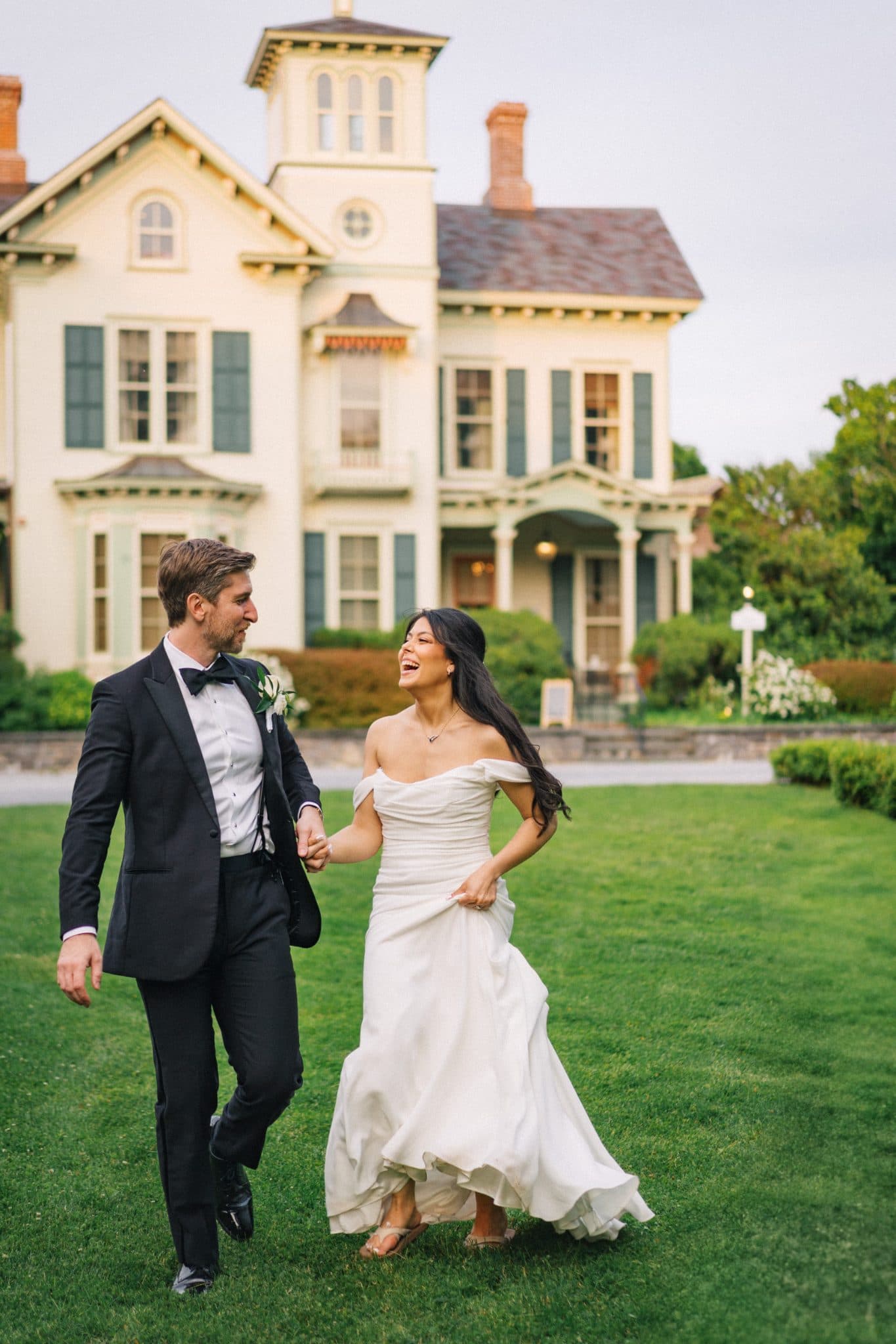 A bride in a white off-shoulder gown and a groom in a black suit walk hand in hand on a grassy lawn in front of a large, multi-story house.