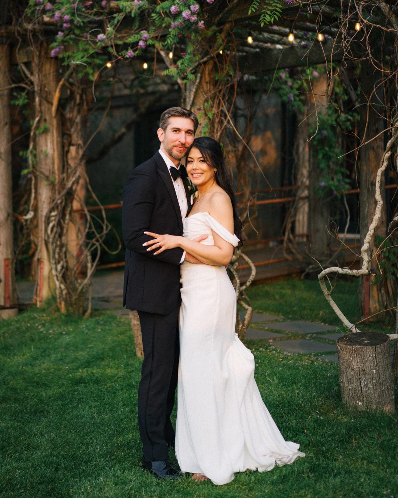 A couple stands together in a garden, the bride in a white dress and the groom in a black suit, under a trellis with purple flowers and string lights.