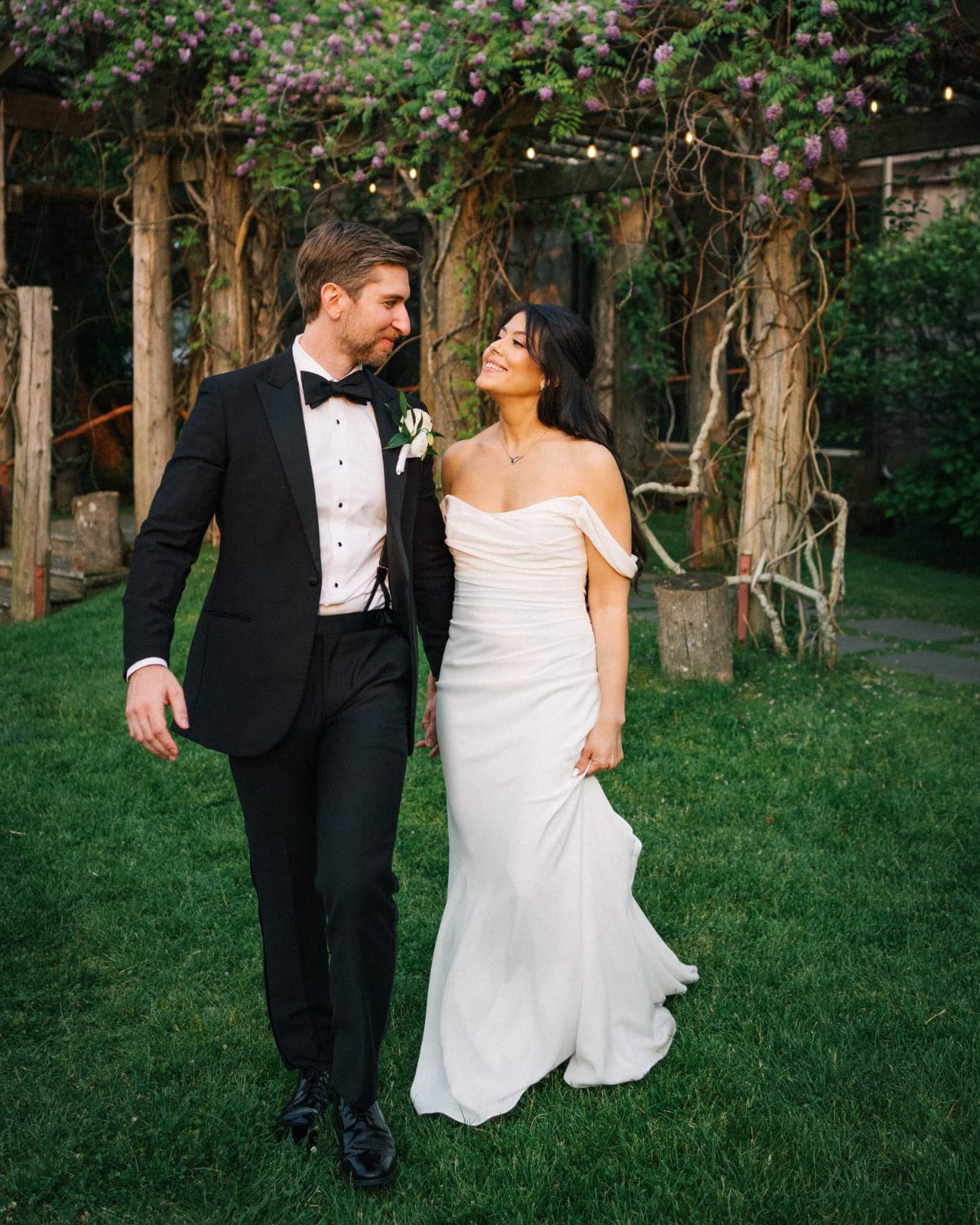 A couple walks hand in hand on grass under a floral archway.