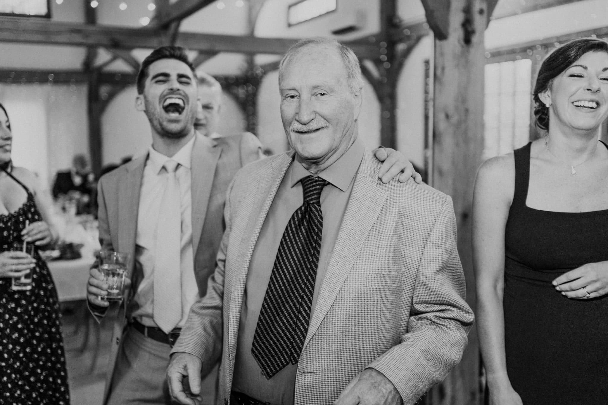 A black-and-white photo captures a group of people laughing and smiling in a warmly lit indoor setting.