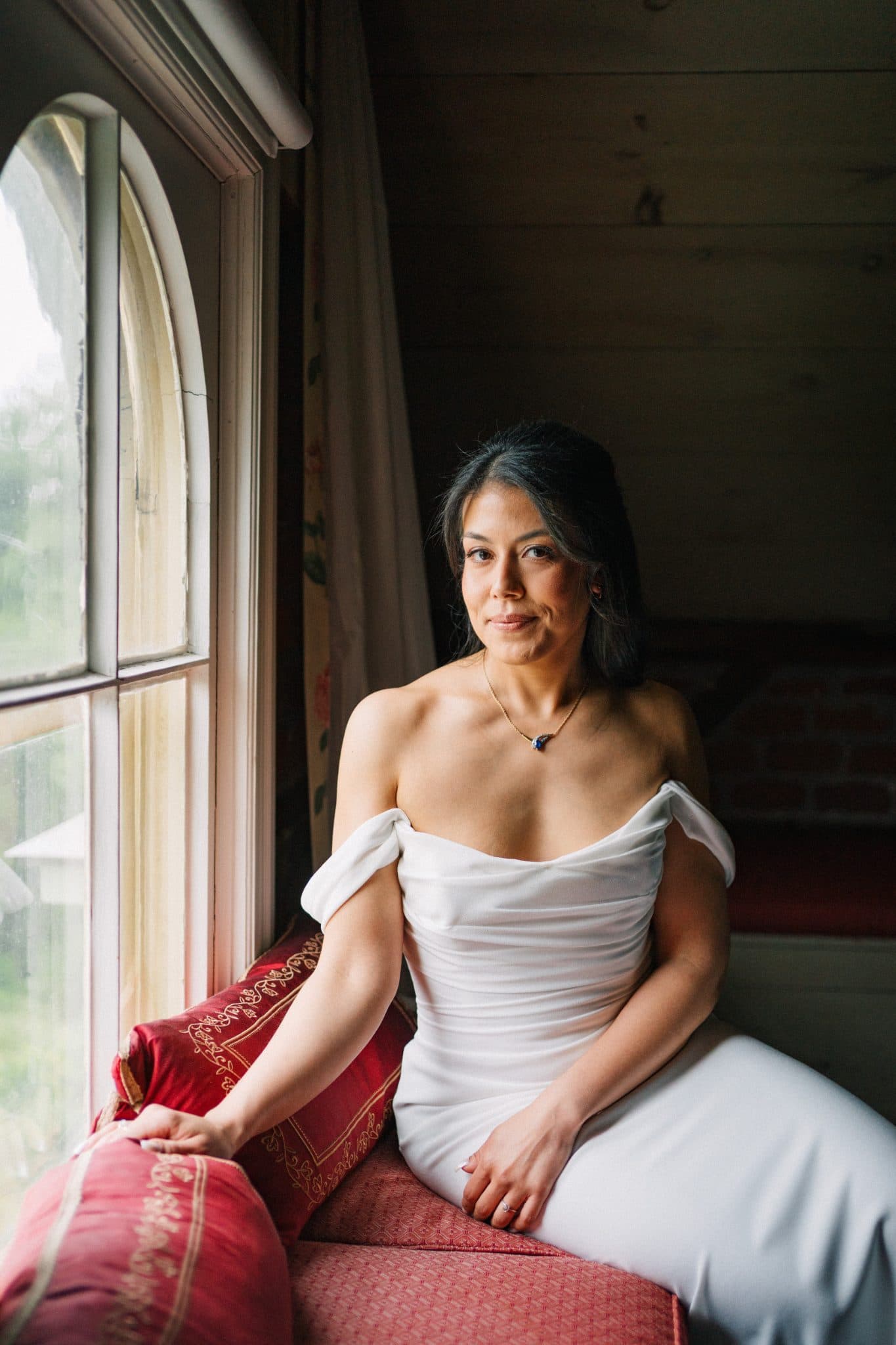 A woman in a white off-shoulder dress sits on a red cushioned bench by a large arched window.