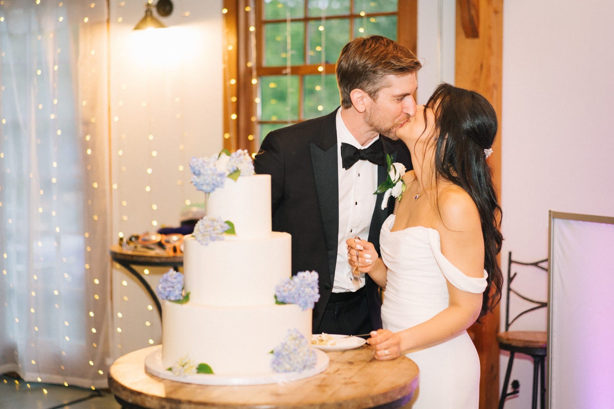 A couple in formal attire stands beside a three-tiered wedding cake adorned with blue flowers, sharing a kiss.