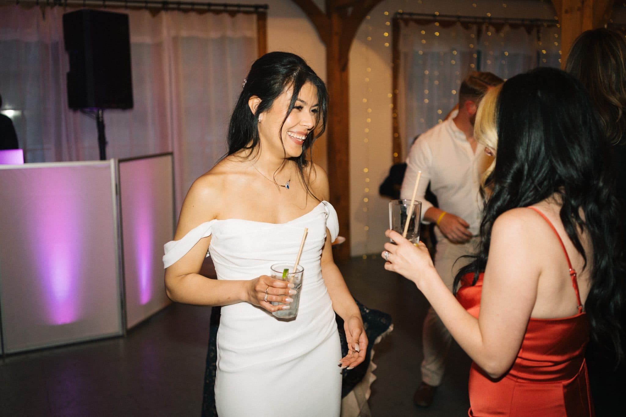 A woman in a white dress holds a drink with a straw, smiling as she interacts with another woman in a red dress at a lively event.