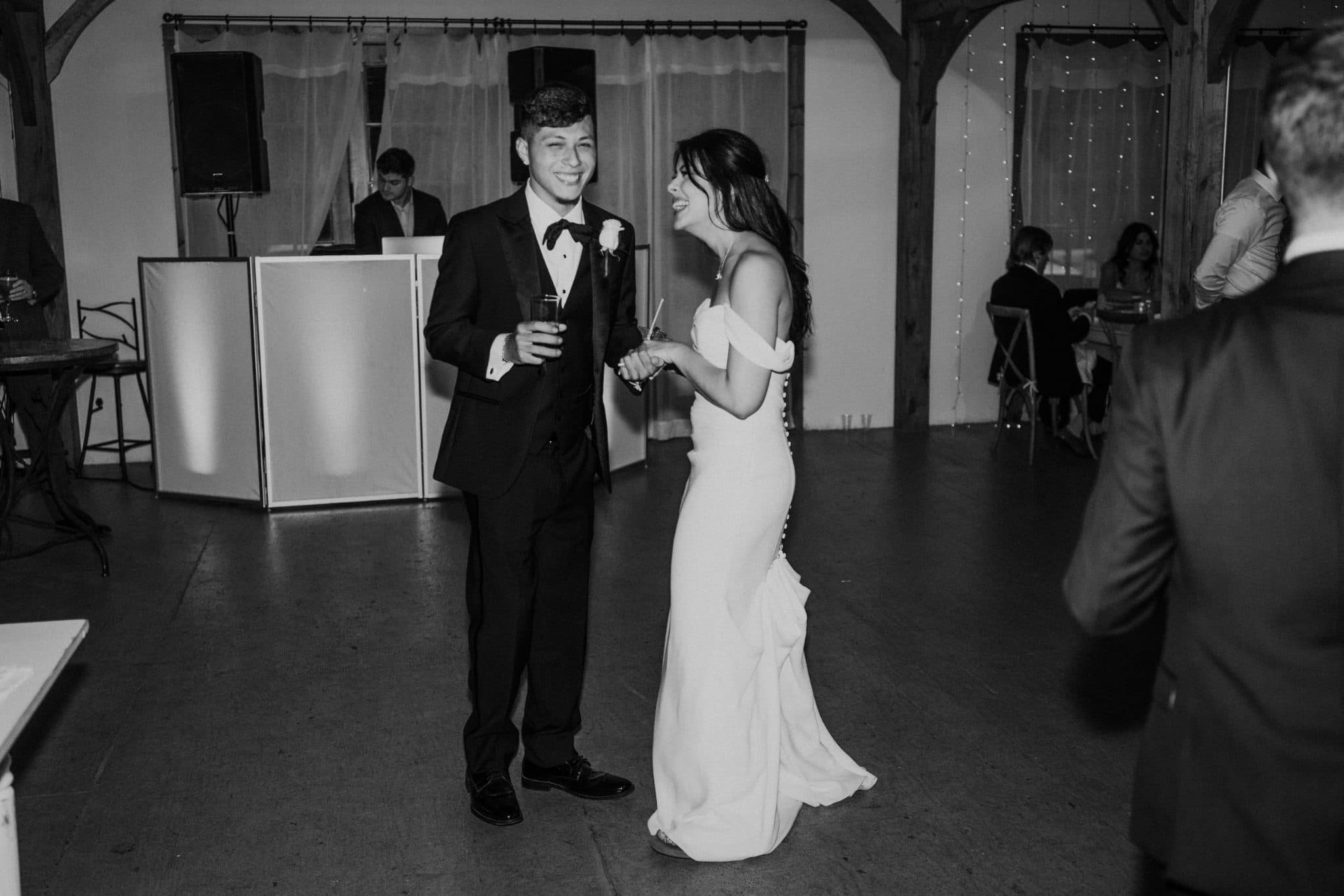 A bride in a white dress and a groom in a black tuxedo hold hands and smile in a wedding reception setting.