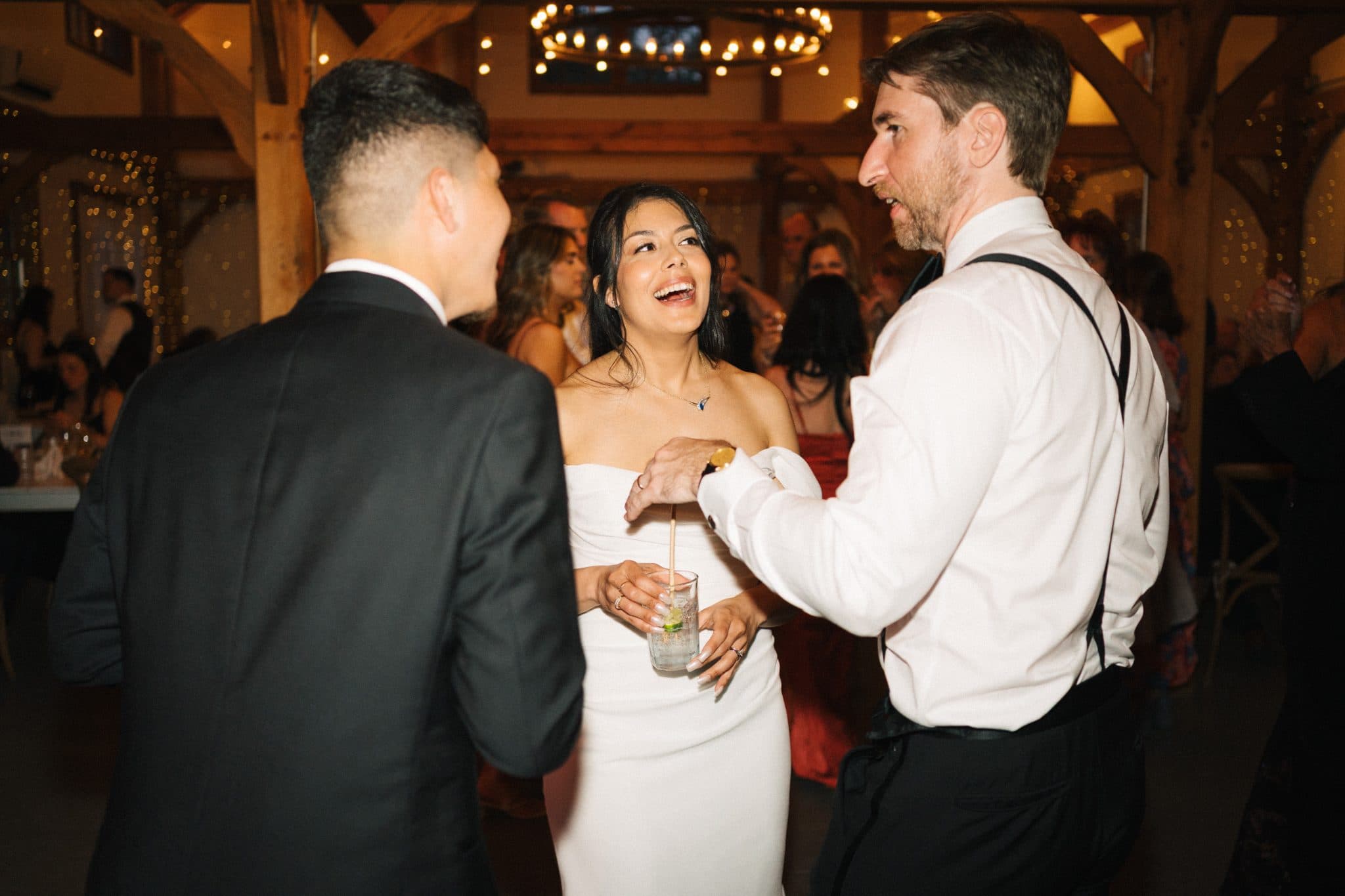 A bride in a strapless white dress laughs while holding a drink, flanked by two men in formal attire at a wedding reception.