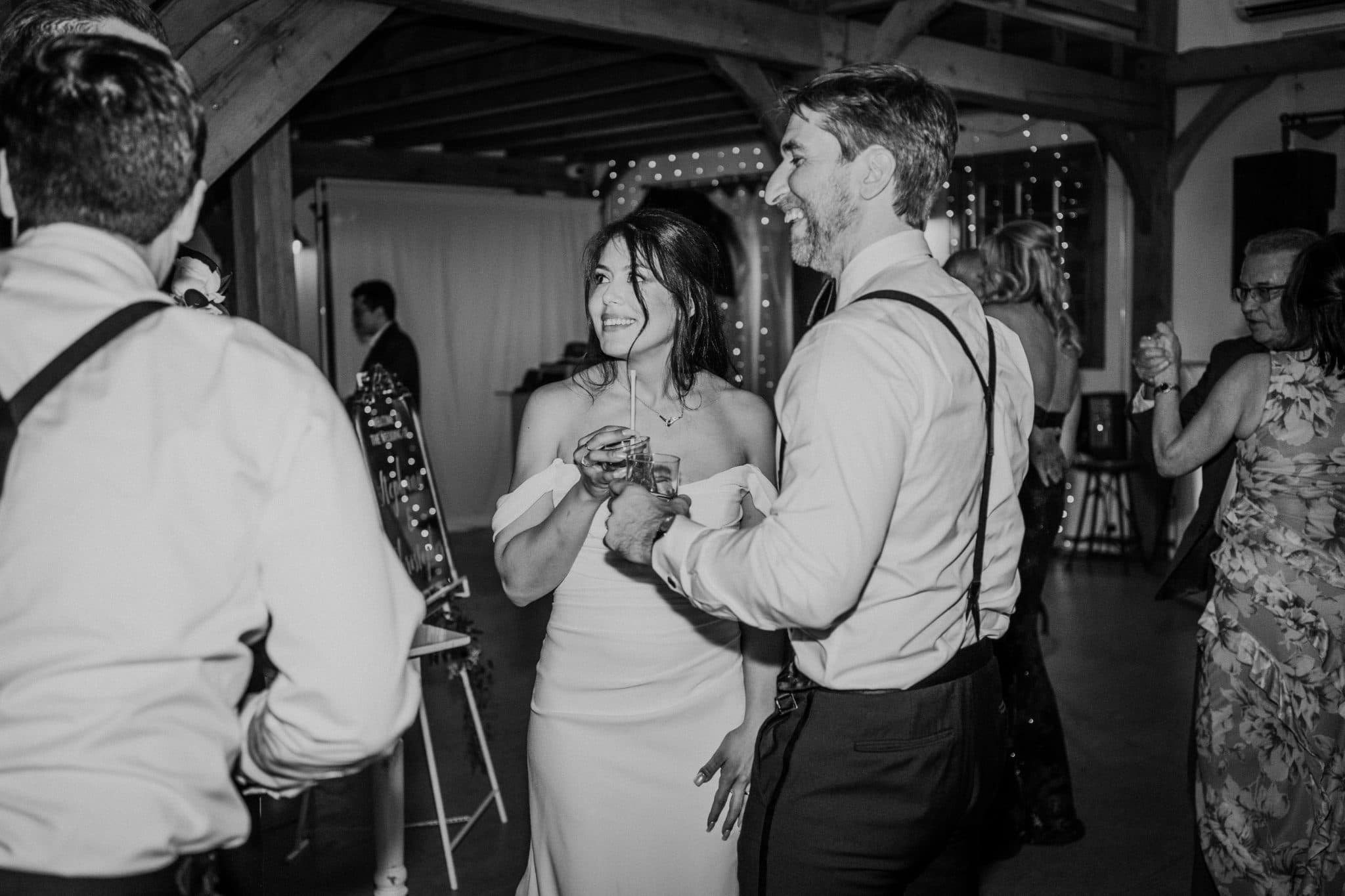 A bride and groom hold drinks and smile at each other during a lively wedding reception.