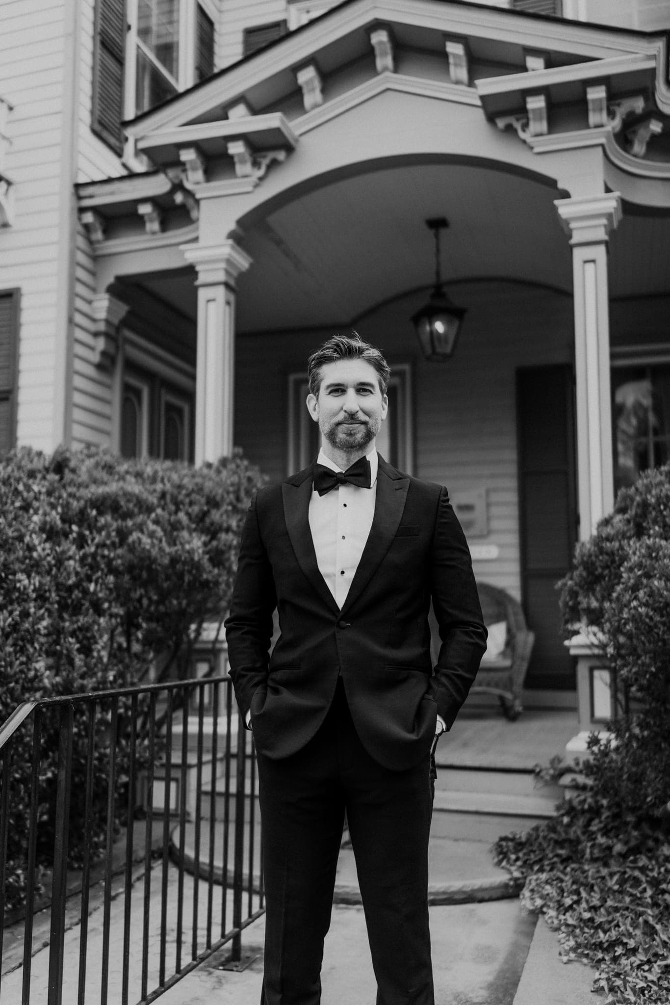 A man in a tuxedo stands in front of a house with a porch and decorative columns.