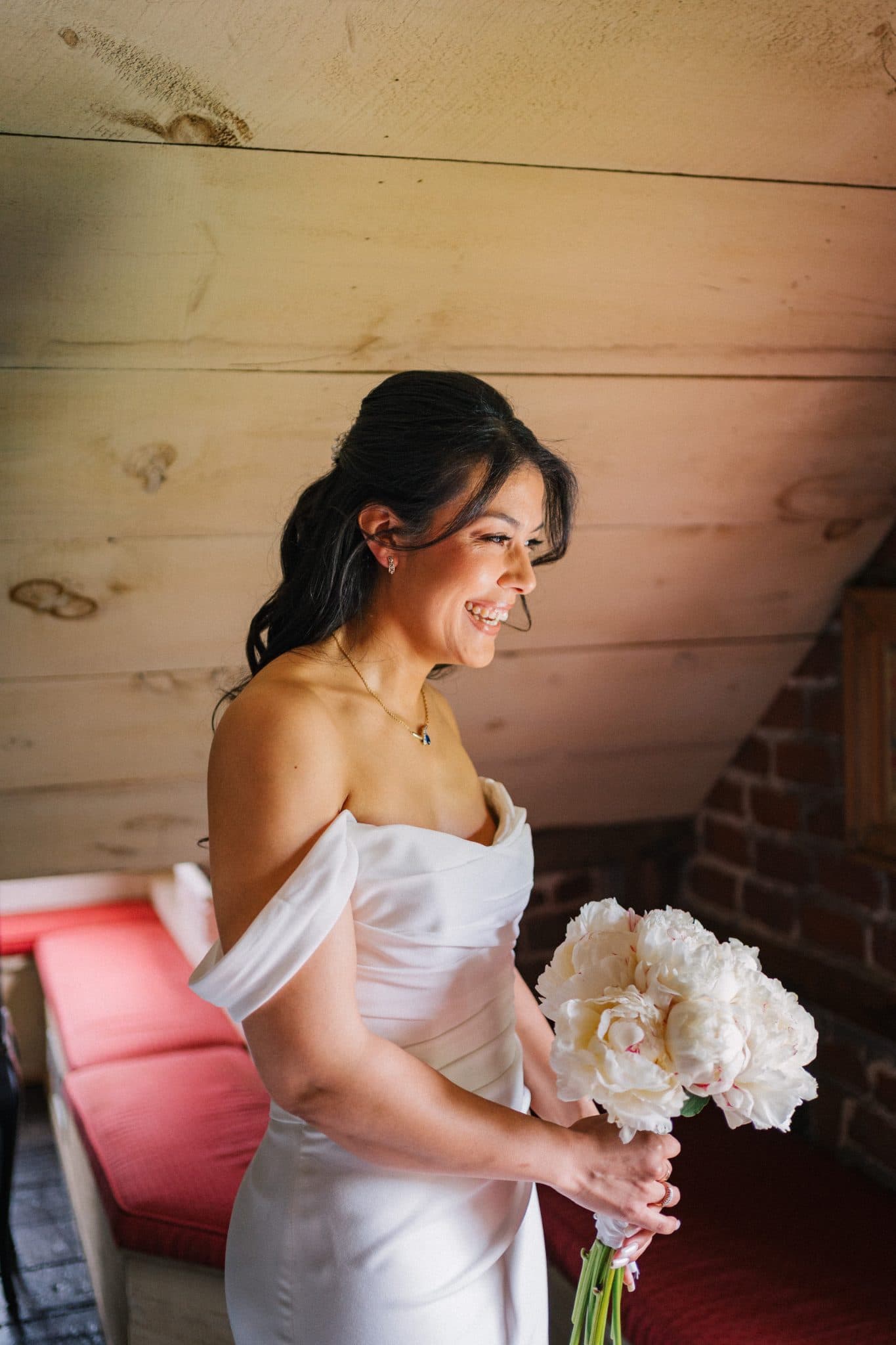 A woman in a white off-shoulder dress holds a bouquet of white flowers, smiling near a wooden wall and red furniture.