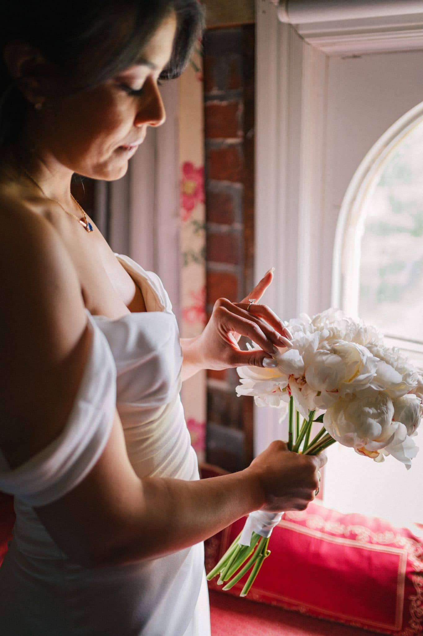A woman in a white dress holds a bouquet of white flowers near a window with red curtains.
