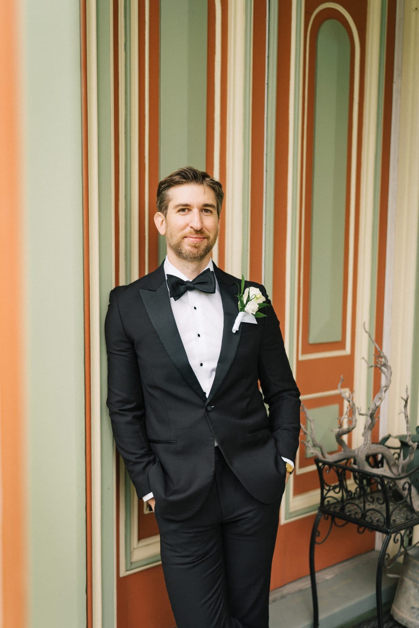 A man in a black tuxedo with a white boutonniere stands in front of a striped wall with a decorative plant nearby.