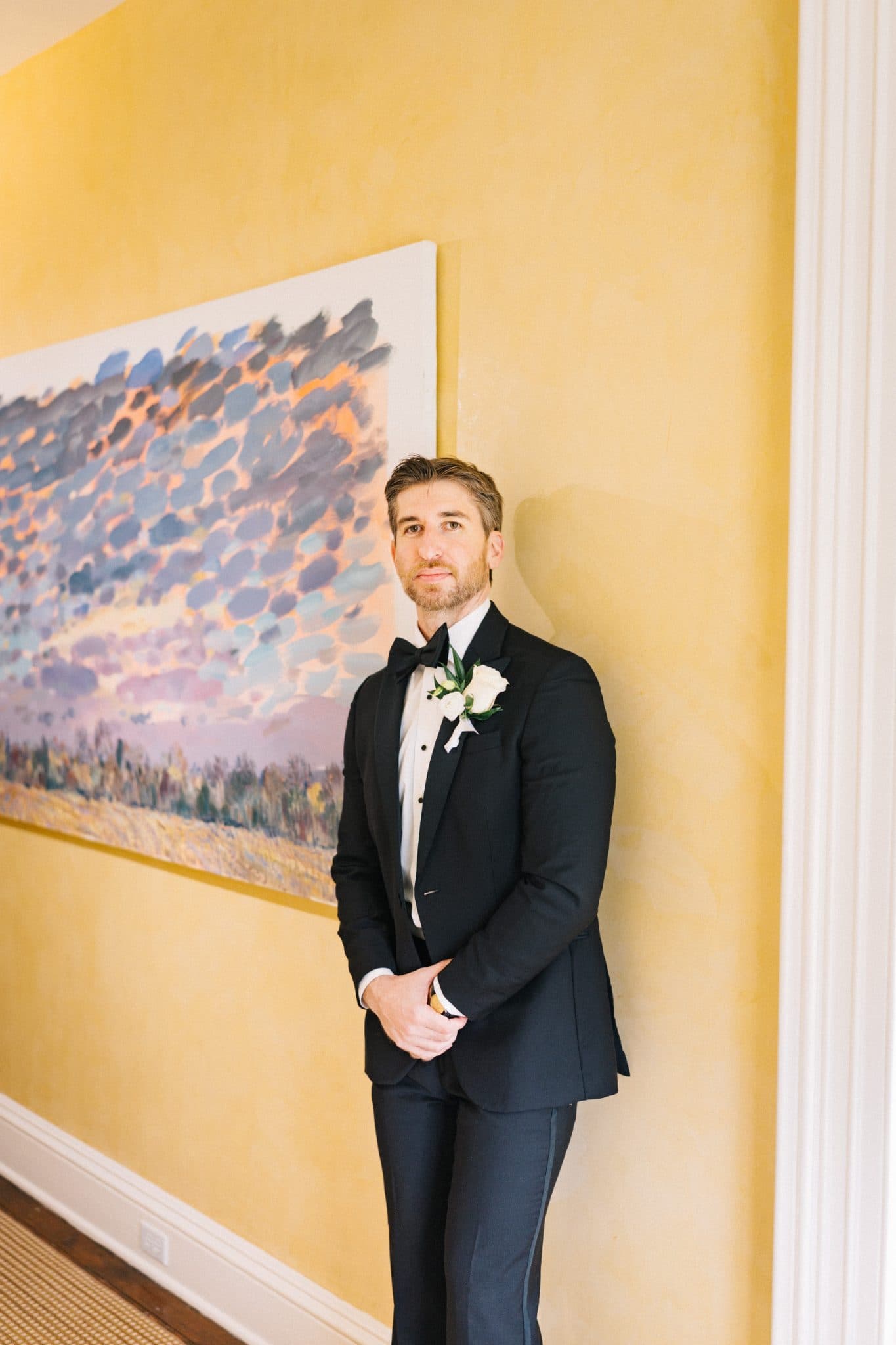 A man in a black tuxedo stands against a yellow wall with an abstract painting behind him.