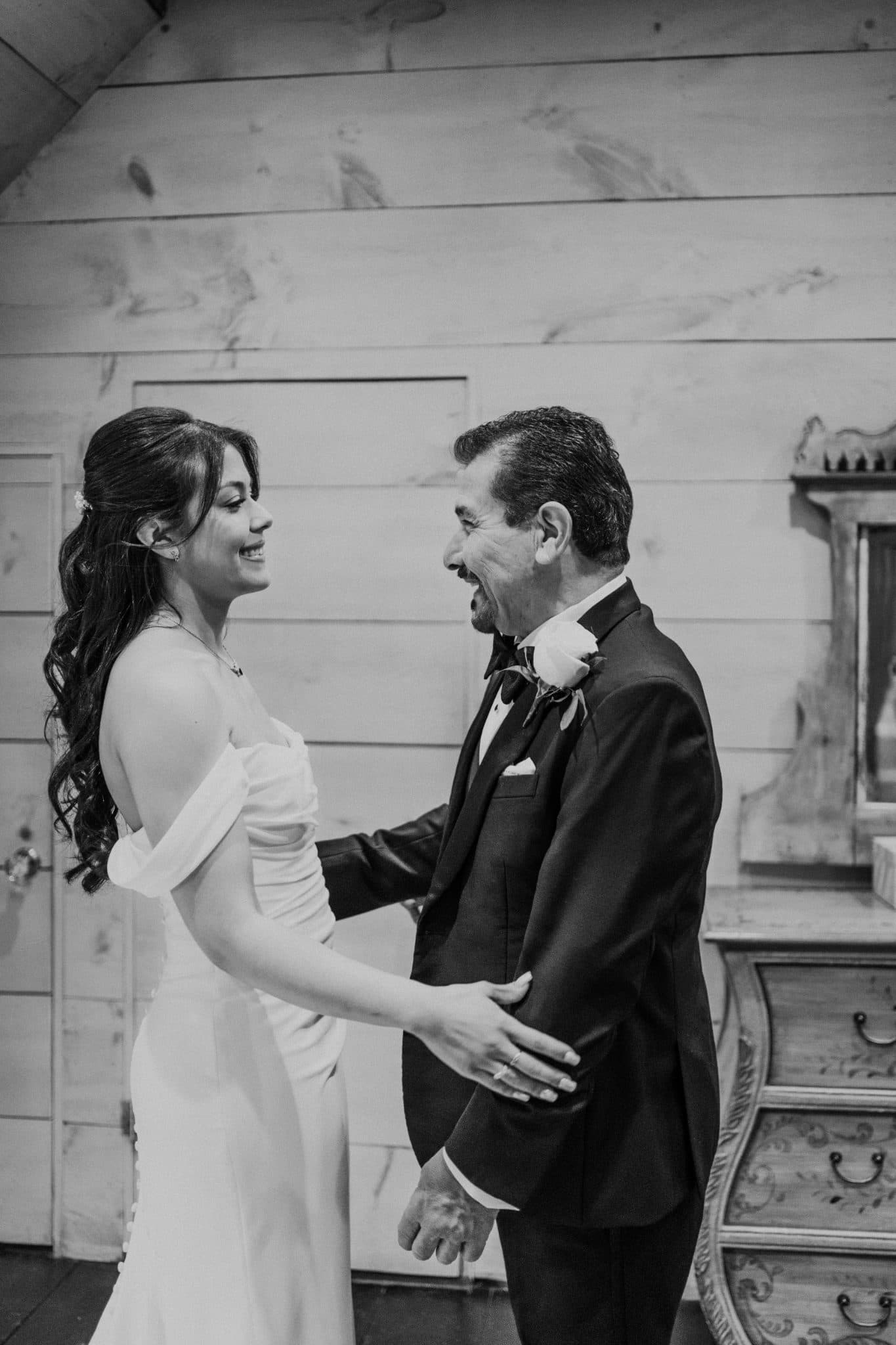 A bride in a white gown and a man in a suit hold hands, smiling at each other.