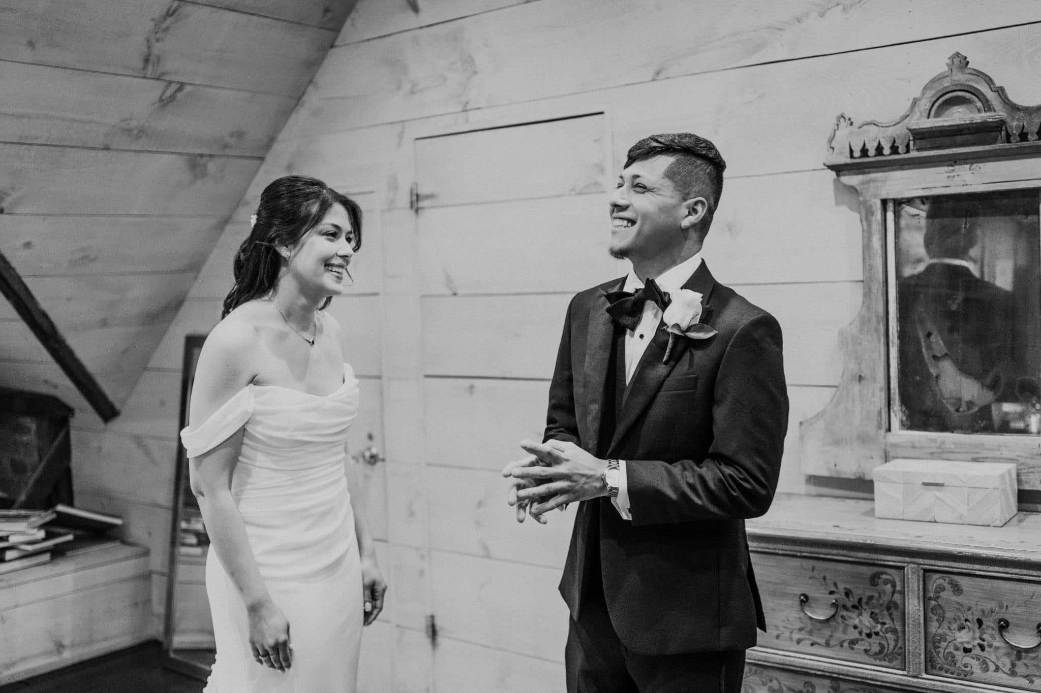 A bride in a strapless white gown and a groom in a black suit with a bow tie stand together, smiling and looking at each other in a rustic setting.
