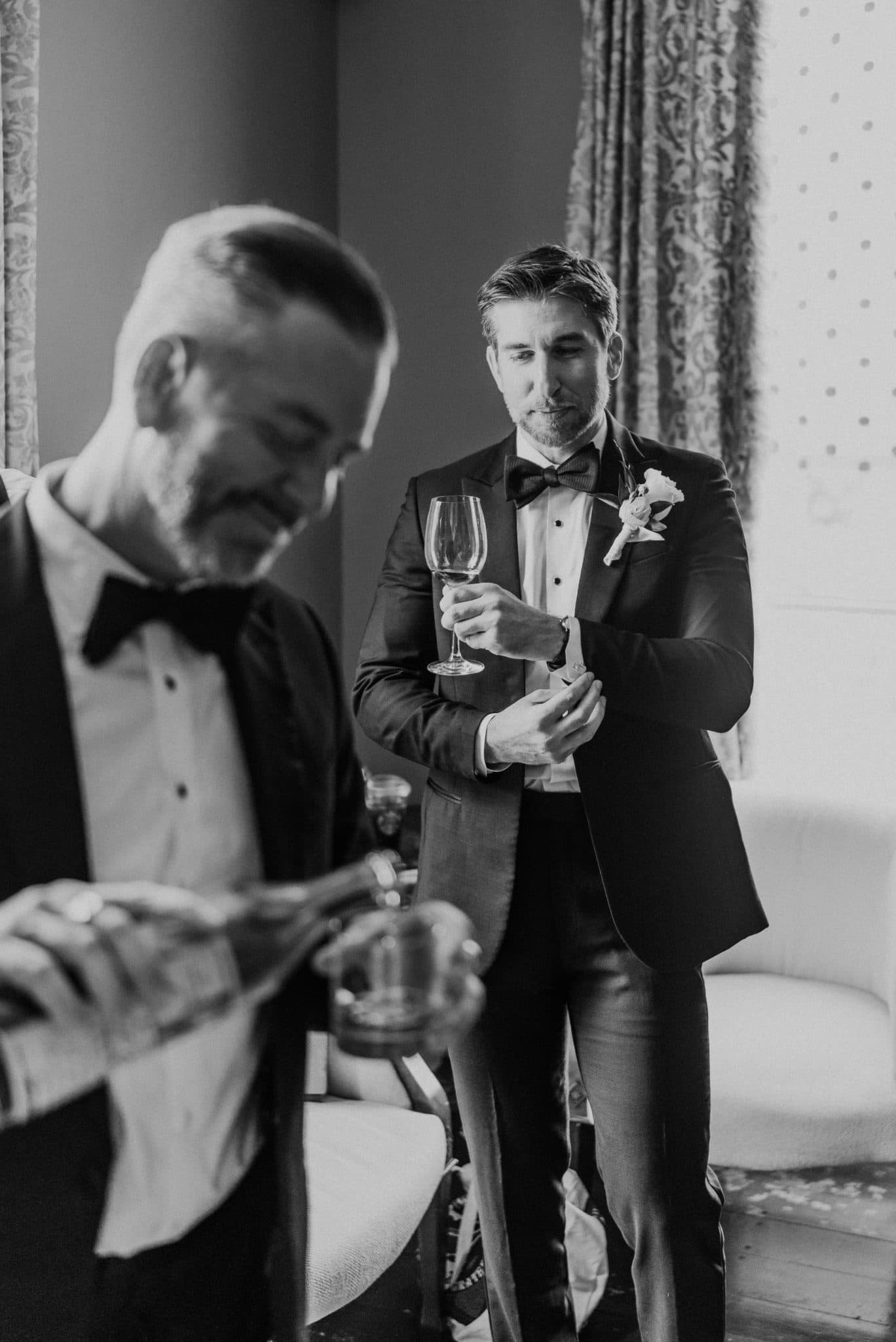 A man in a tuxedo adjusts his boutonniere while another man pours champagne in a formal setting.