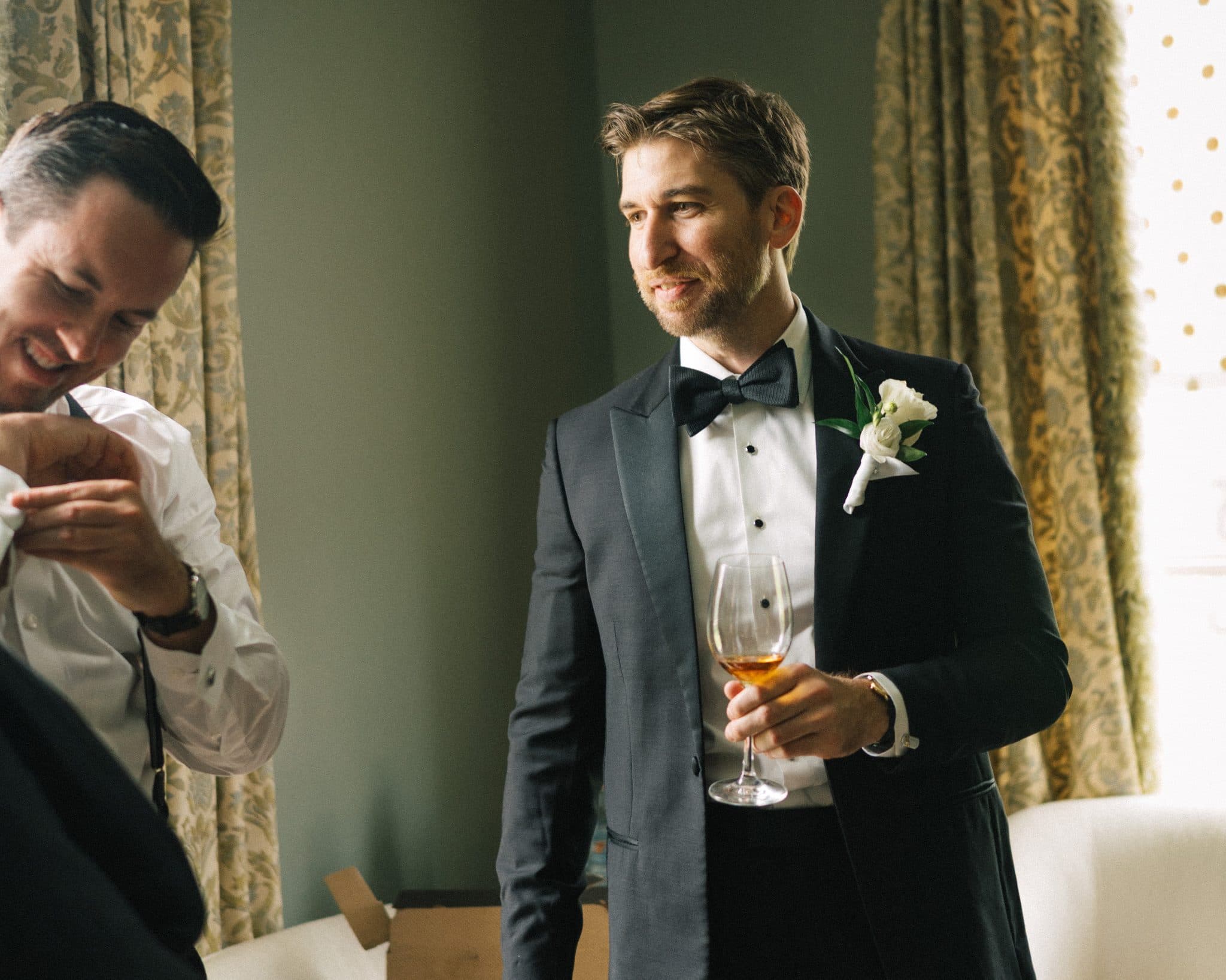 A man in a tuxedo holds a glass of amber liquid, smiling, while another adjusts his shirt in the background.