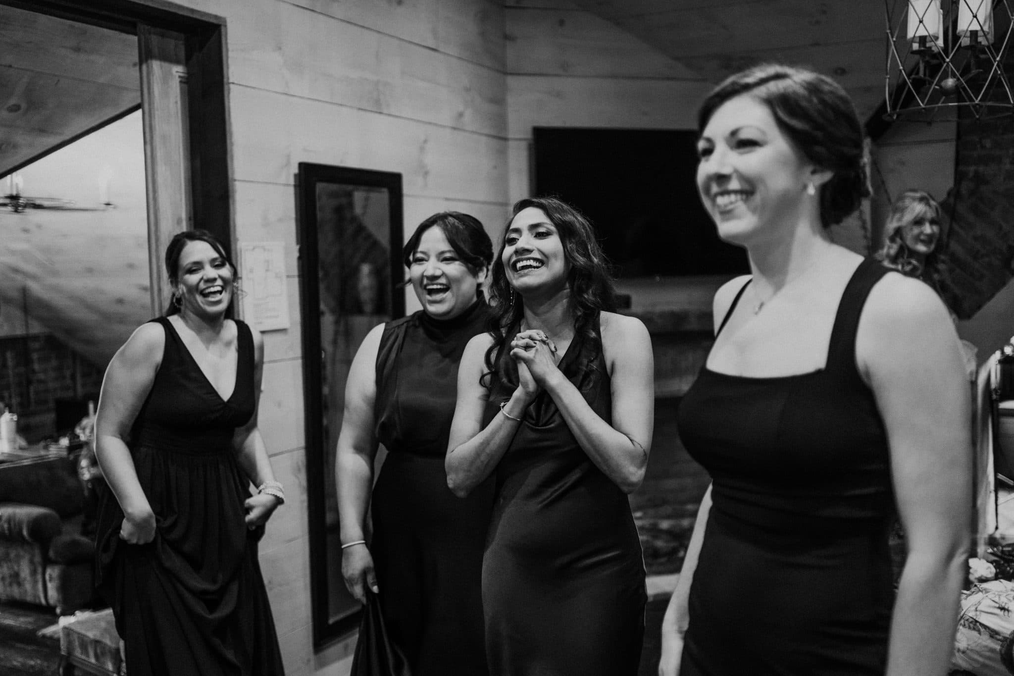 Four women in black dresses stand together, laughing and smiling in a room with wooden walls and framed artwork.