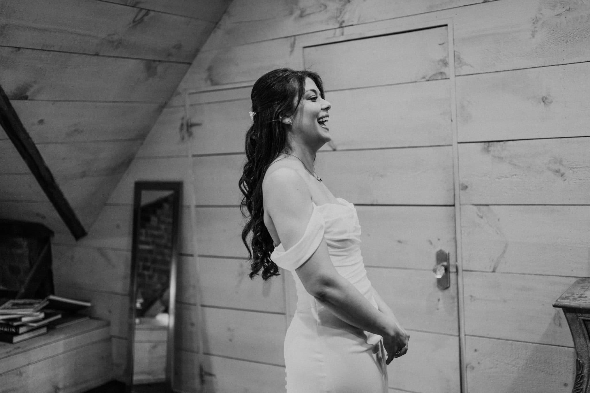 A bride in a white dress stands in a rustic room, smiling and adjusting her dress.