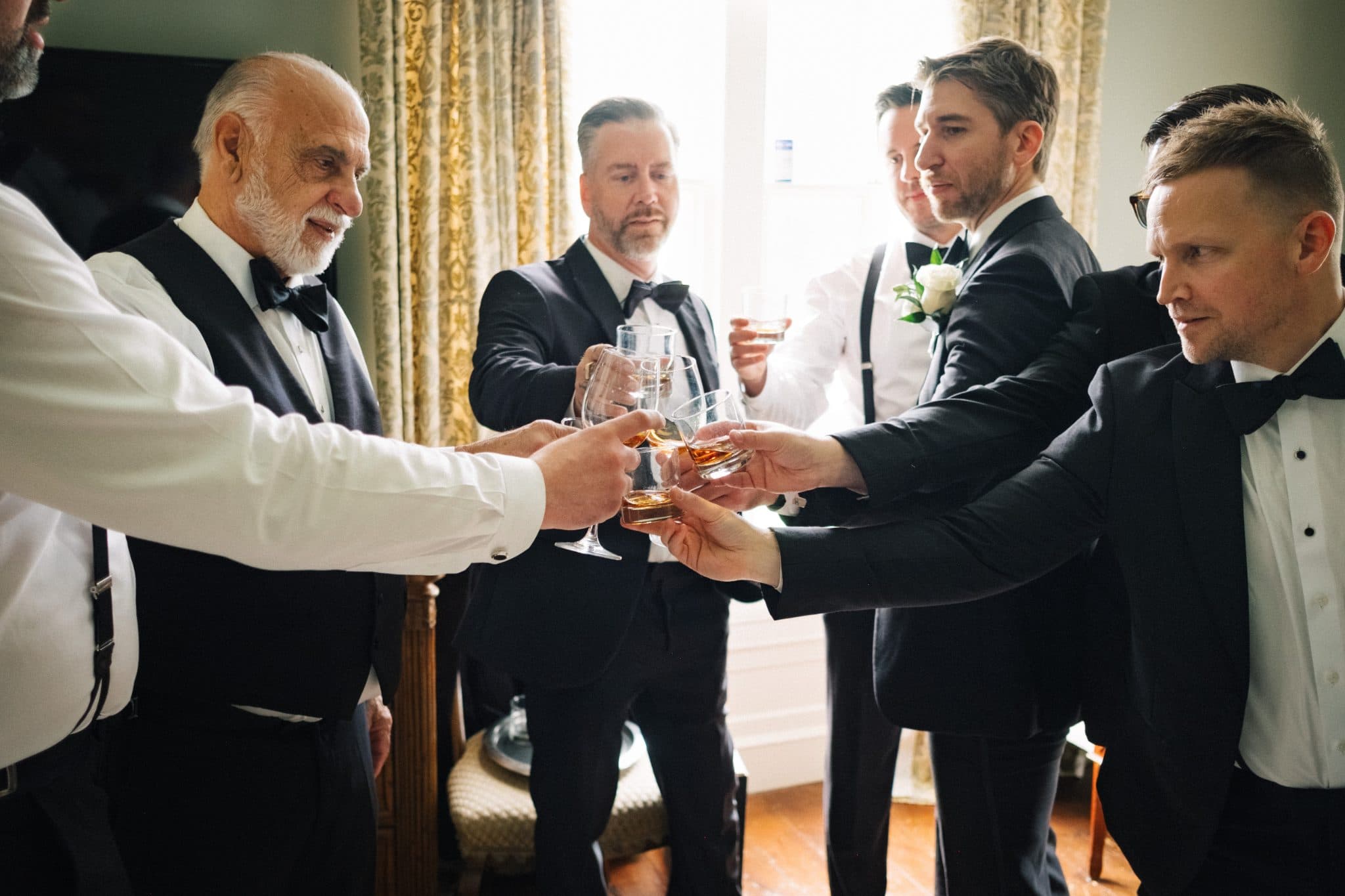 A group of men in formal attire toasts with glasses of whiskey in a warmly lit room.