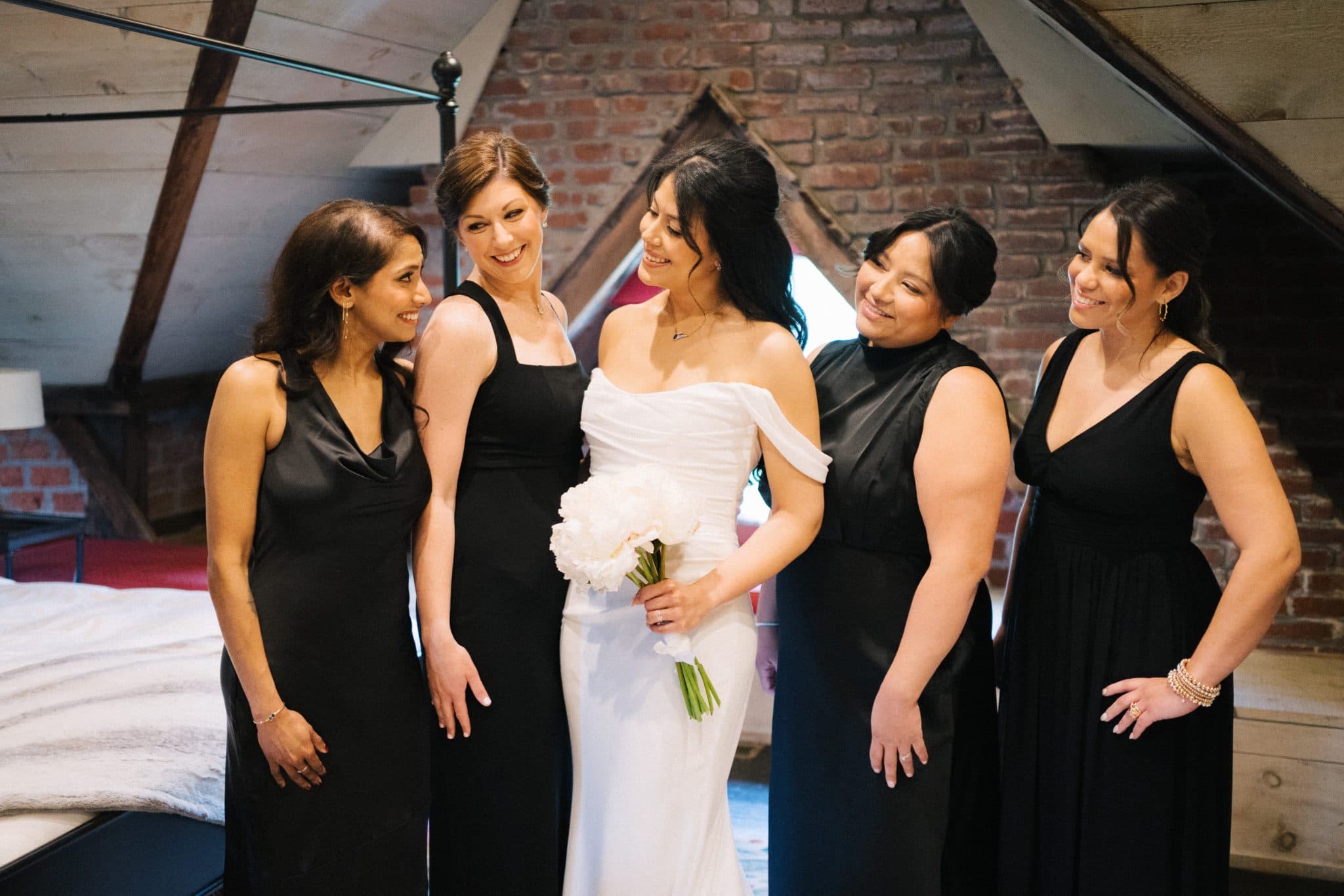 A bride in a white dress holds a bouquet, surrounded by four women in black dresses, smiling in a room with a brick wall and a bed.