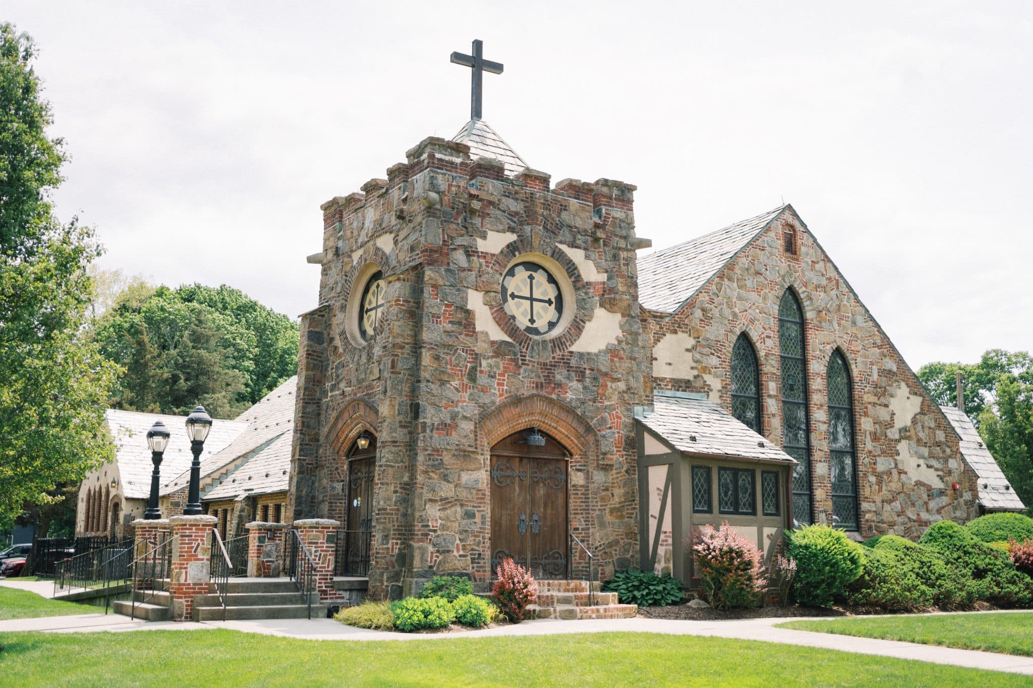 A stone church with a cross on top, surrounded by greenery and a sidewalk.