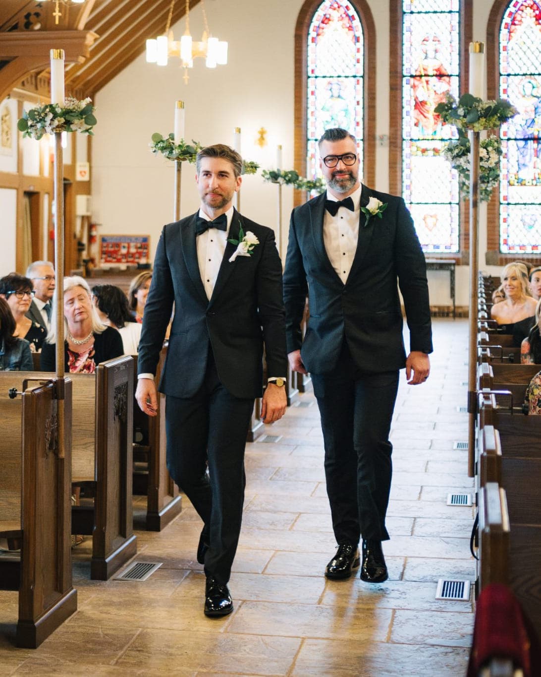 Two men in suits walk down a church aisle, surrounded by seated guests.