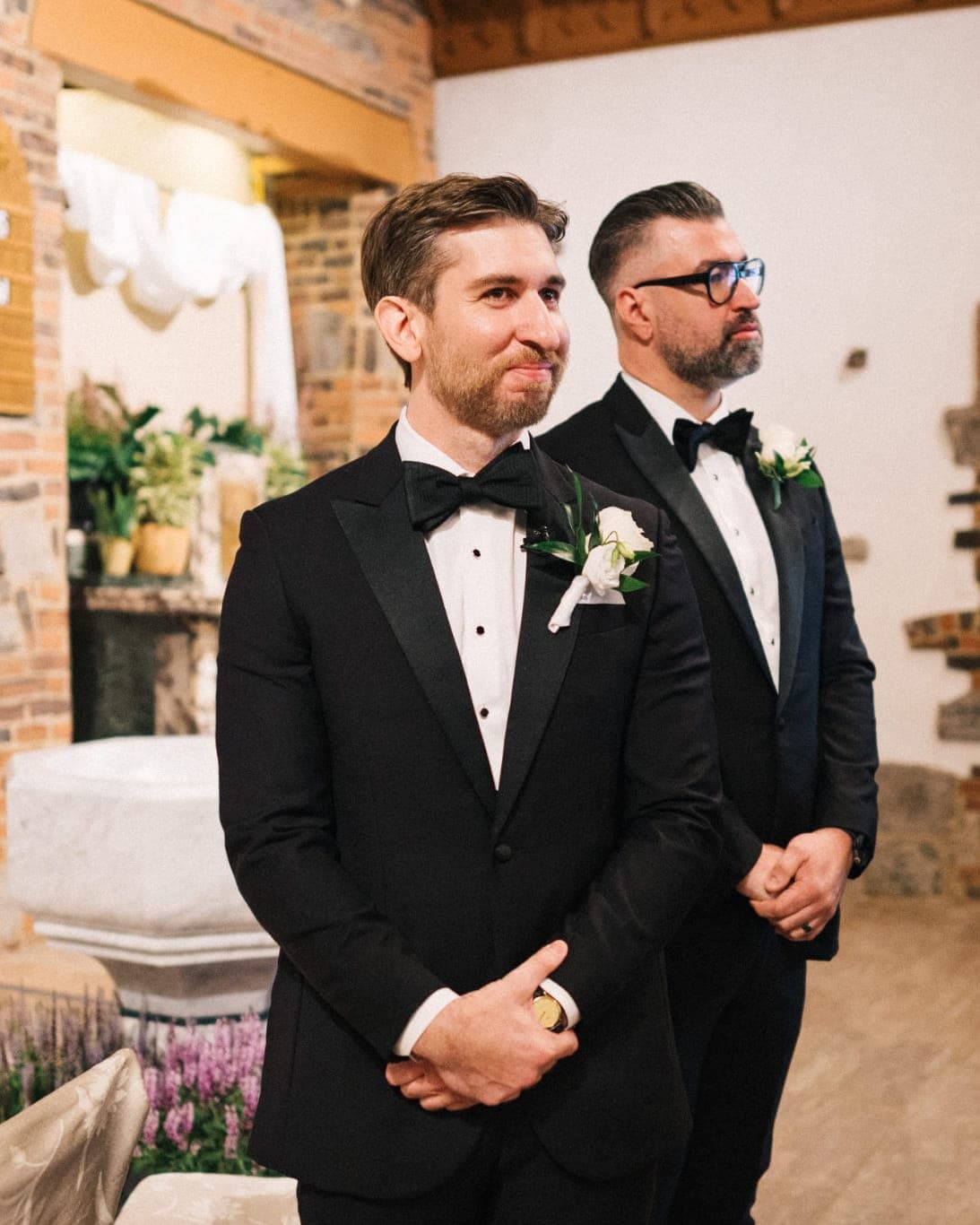 Two men in black tuxedos stand in a church, one with a boutonniere, both looking forward.