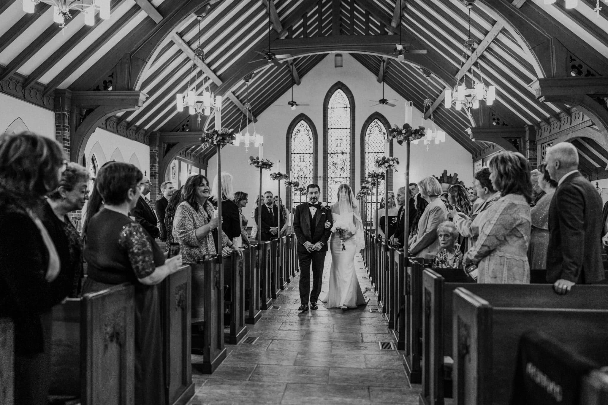 A bride and groom walk down the aisle in a church, surrounded by guests seated in pews.