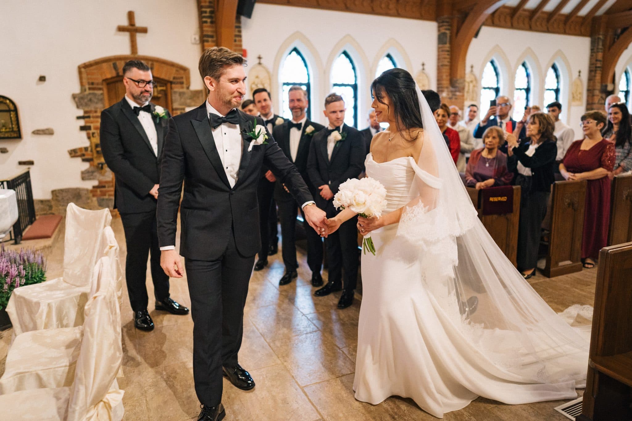 A bride in a white dress and a groom in a black tuxedo hold hands as they walk down the aisle in a church.