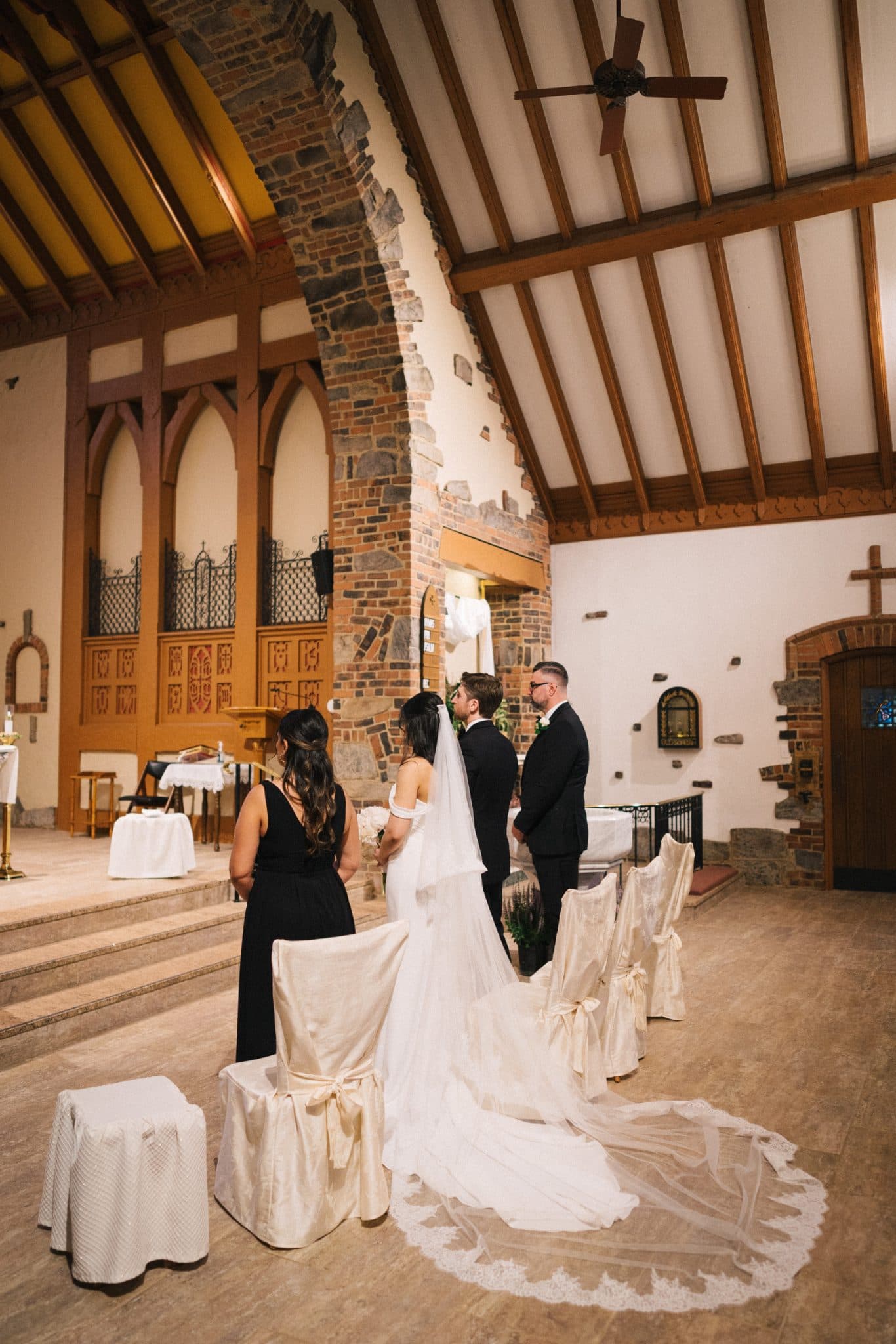 A bride and groom stand at the altar in a church, facing each other, with a guest in black attire standing nearby.