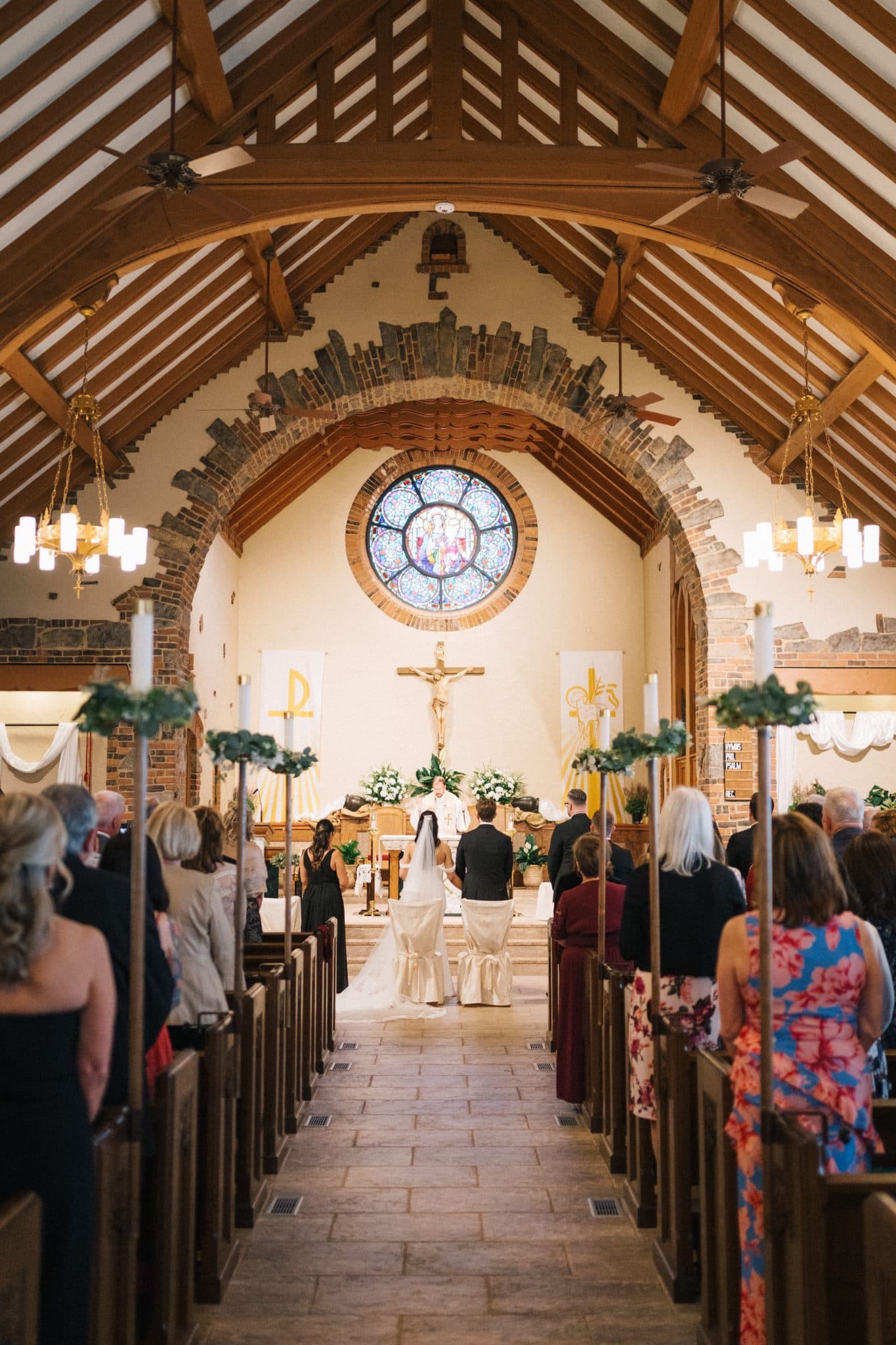 A bride and groom stand at the altar in a church with wooden beams, a stained glass window, and floral decorations.