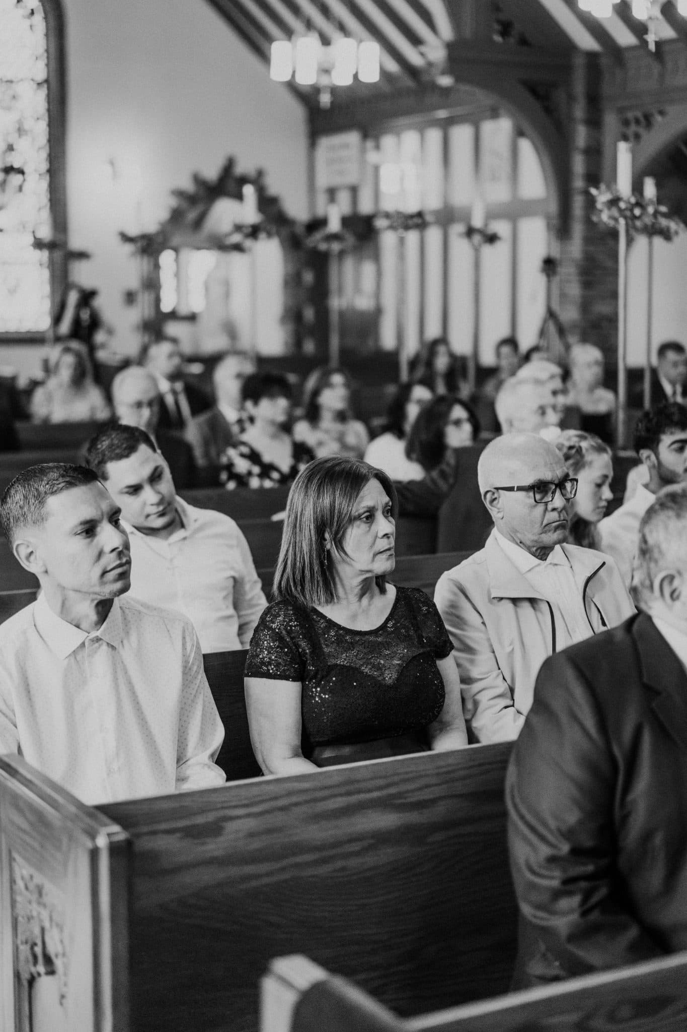 A black and white photo shows a church interior with people seated in pews, some facing forward and others looking to the side.