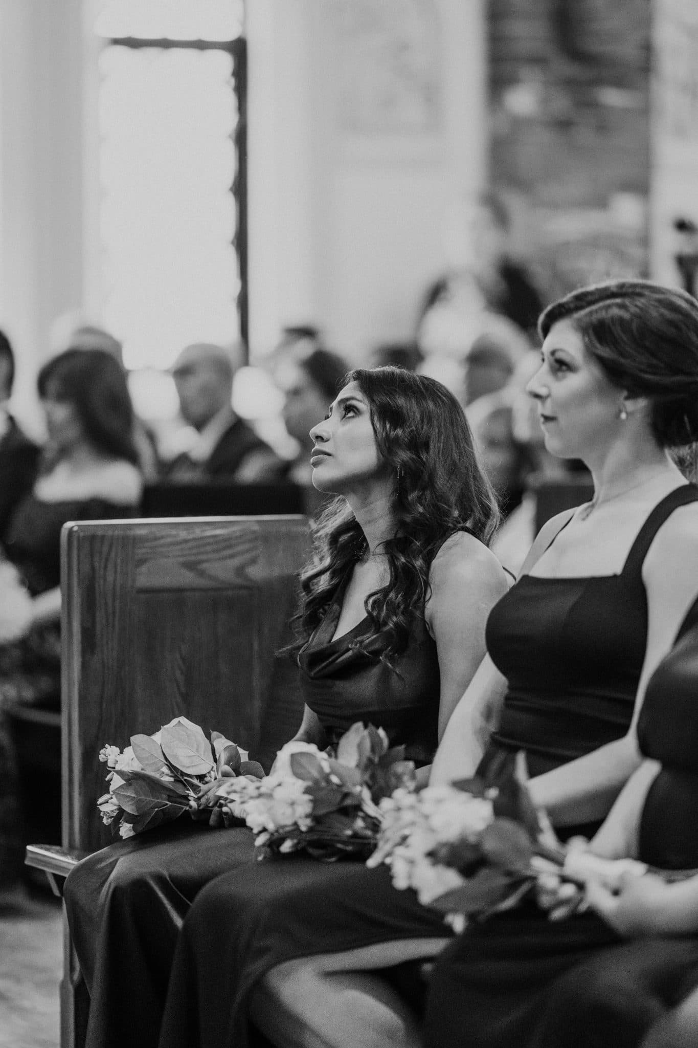 Two women in dark dresses hold bouquets, seated on wooden pews in a church.