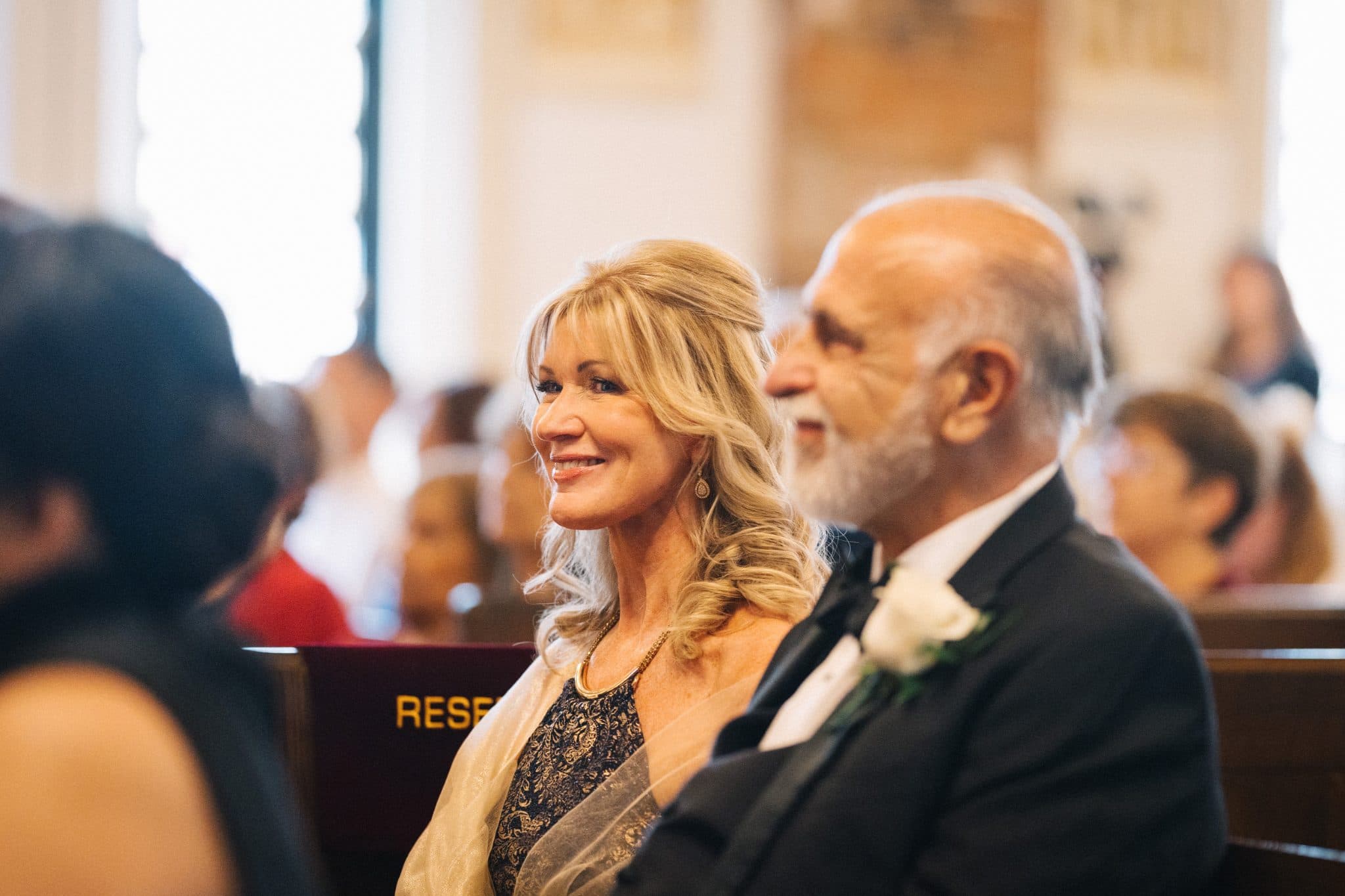 A woman in a lace dress and a man in a suit with a white flower on his lapel sit together in a church pew, smiling.