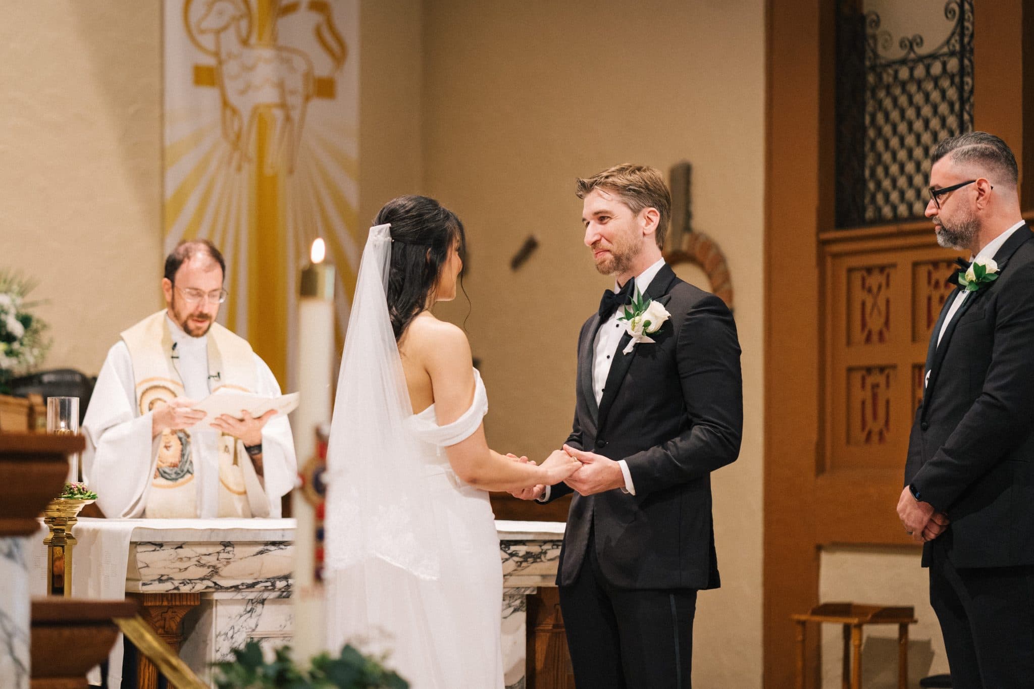 A bride and groom hold hands at a wedding ceremony, with a priest and another man standing nearby.