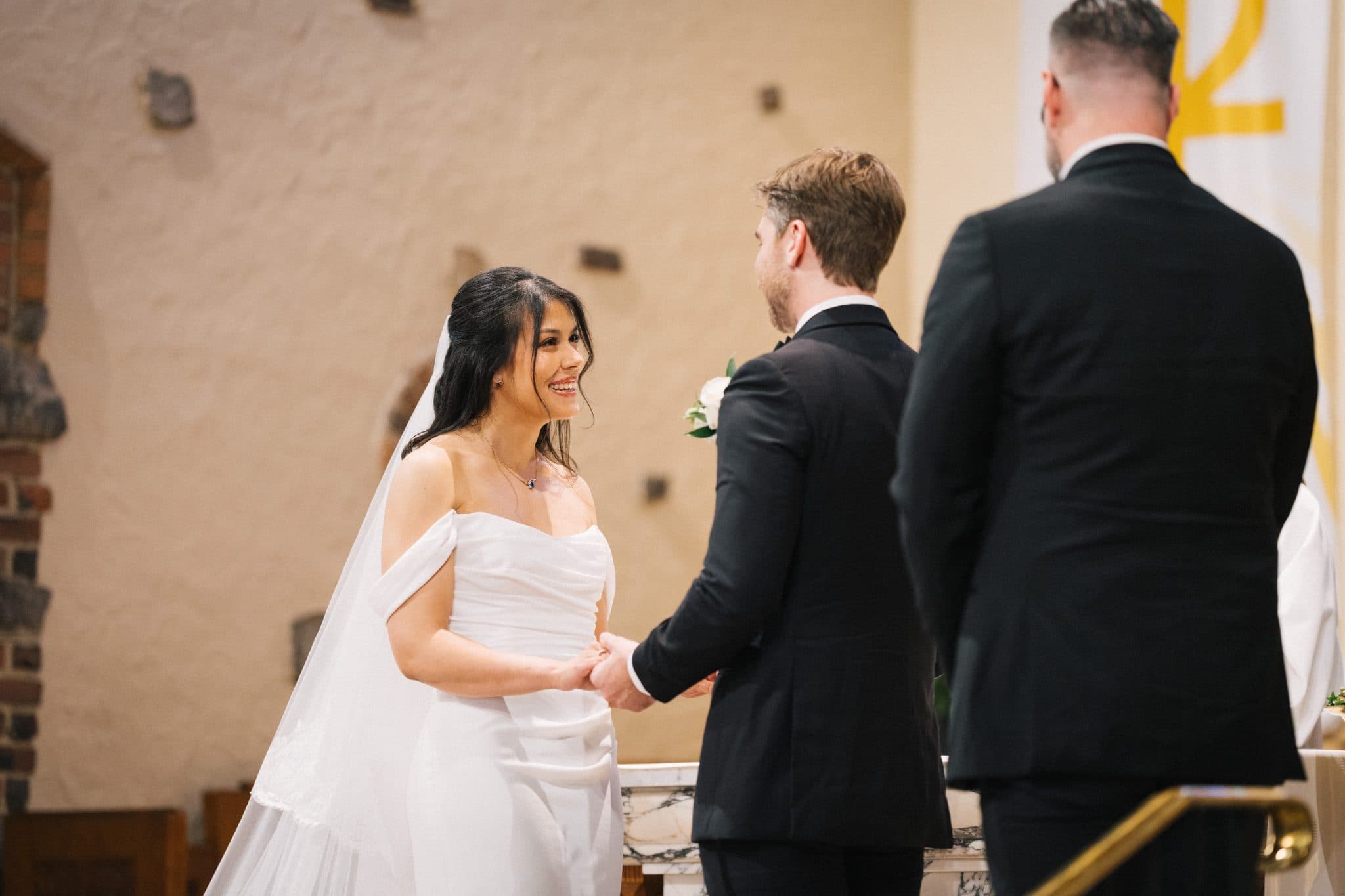 A bride in a white off-shoulder gown and veil holds hands with a groom in a black suit, standing in a church setting.