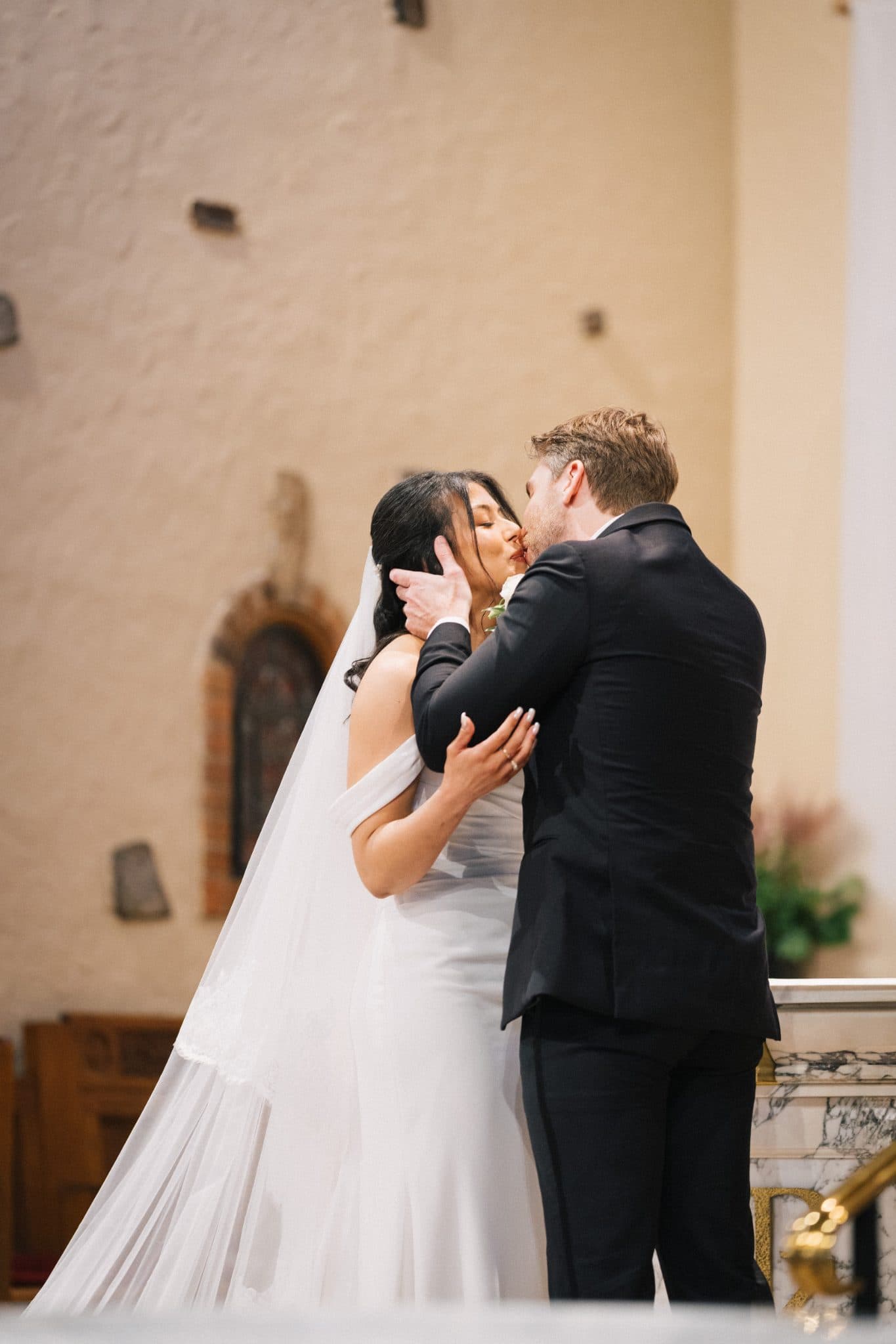 A bride in a white dress and veil kisses a groom in a black suit in a church setting.