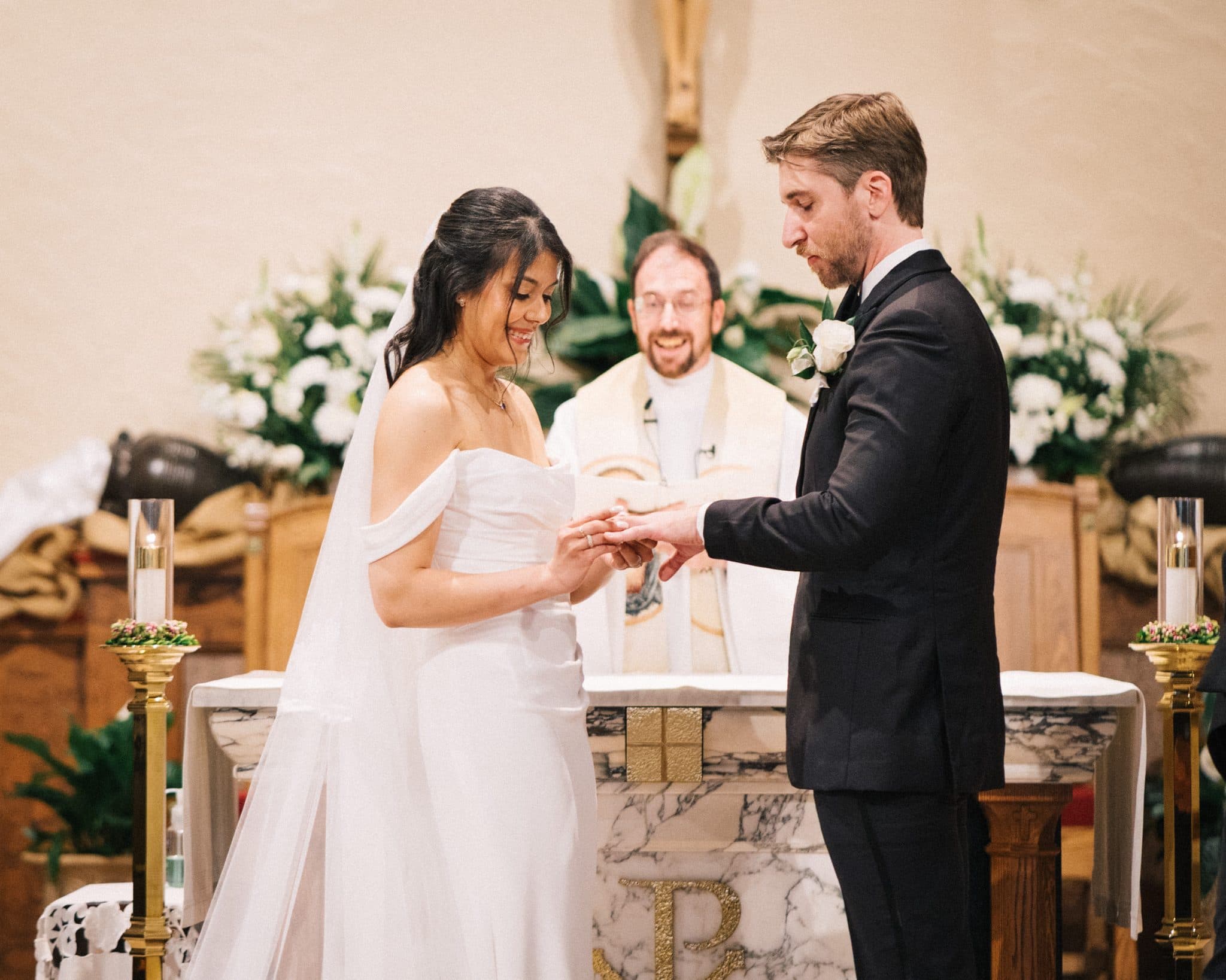 A bride in a white dress and veil exchanges rings with a groom in a black suit, standing at an altar with a priest and floral decorations.