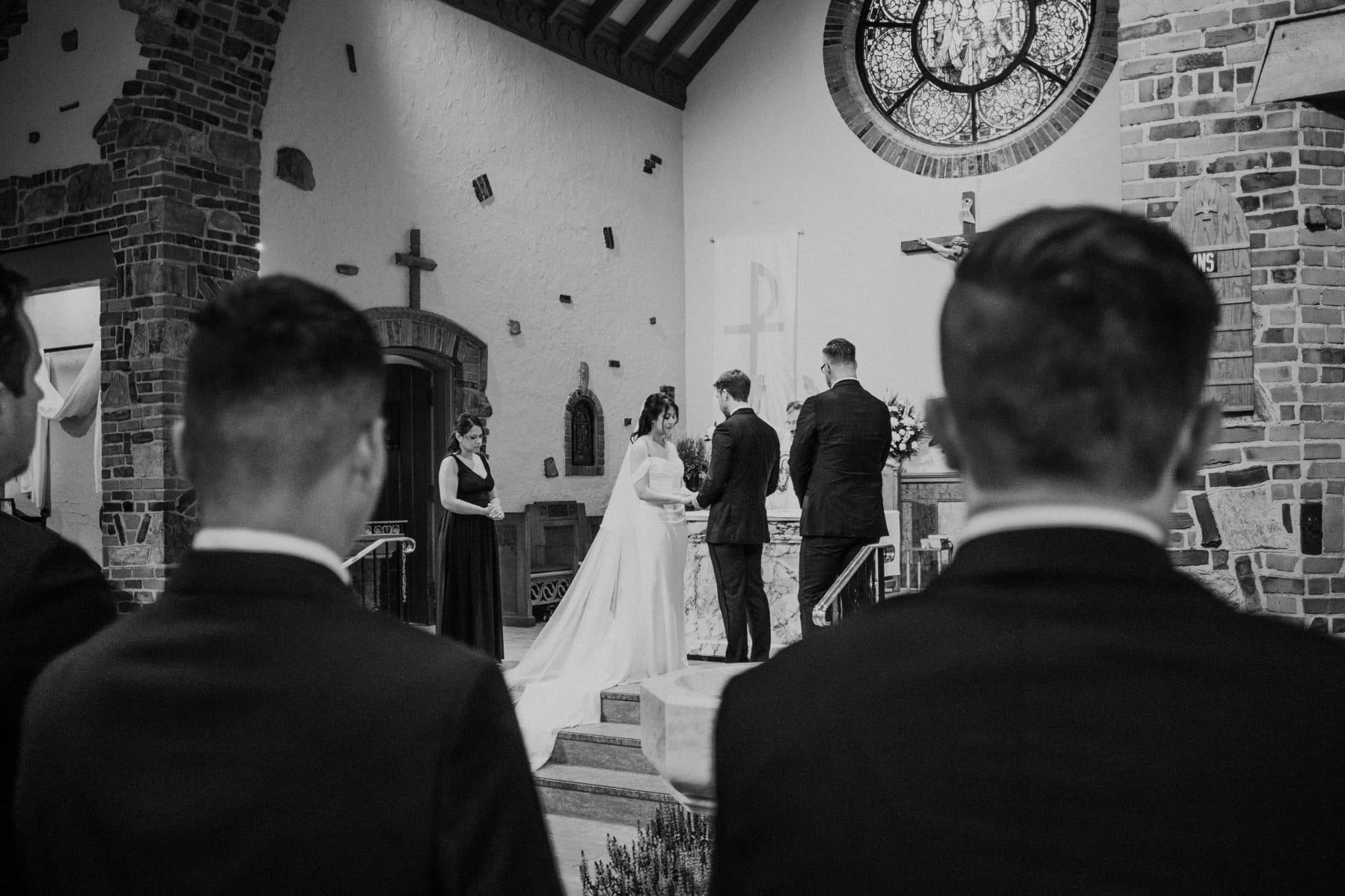 A bride and groom stand at the altar in a church, surrounded by guests and religious symbols.