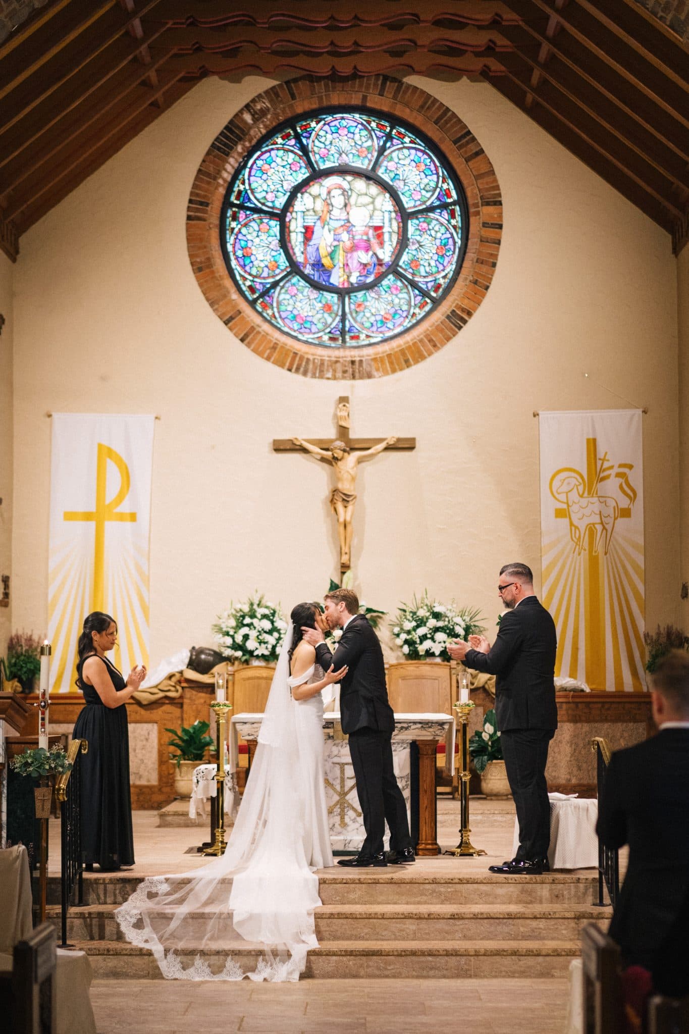 A bride and groom kiss at the altar in a church with a stained glass window and crucifix above.