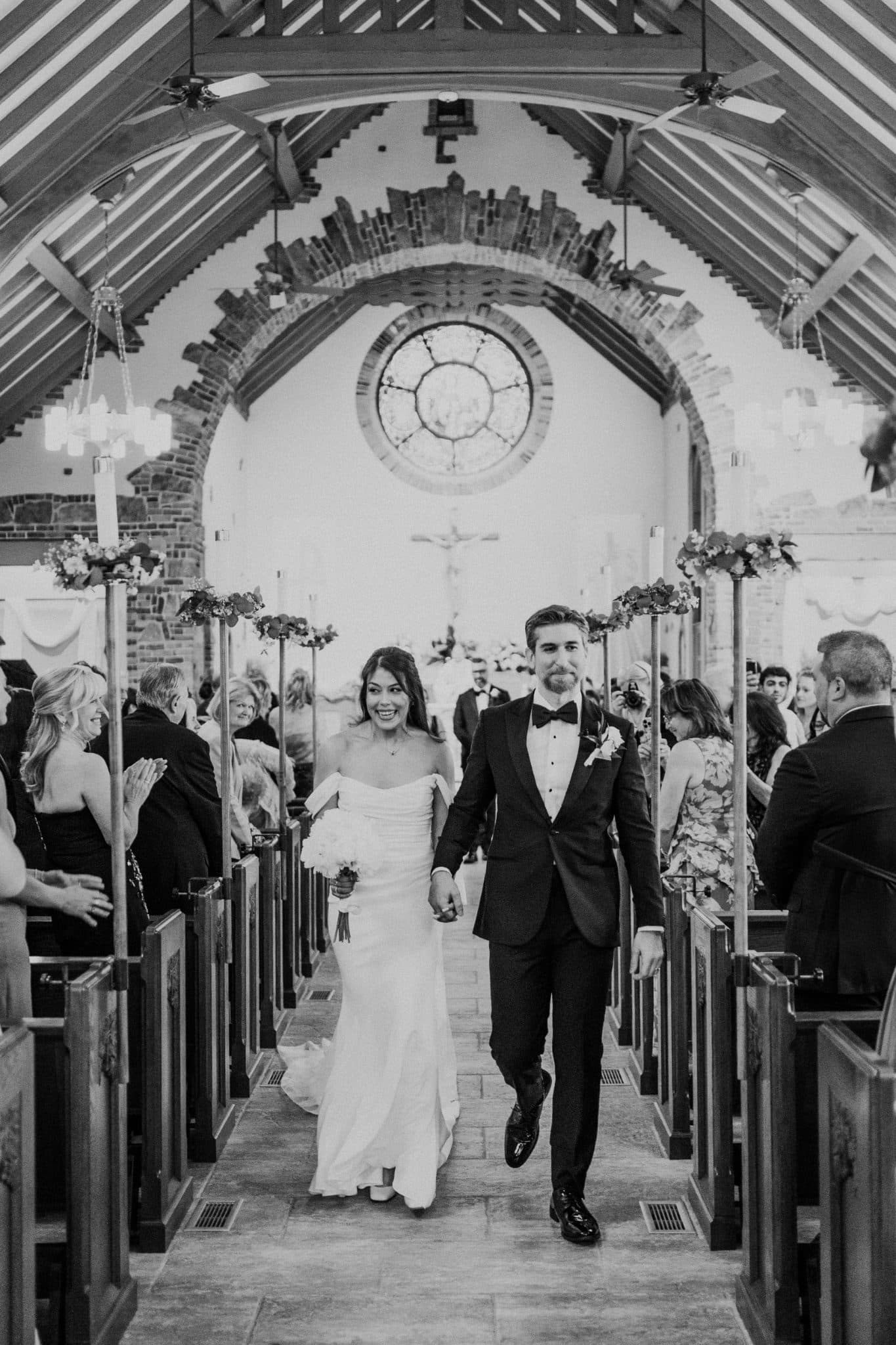 A bride and groom walk down the aisle of a church, holding hands, surrounded by guests.